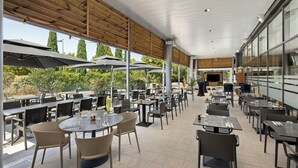 An outdoor dining area with multiple tables and chairs, large umbrellas, and a view of trees and buildings in the background.
