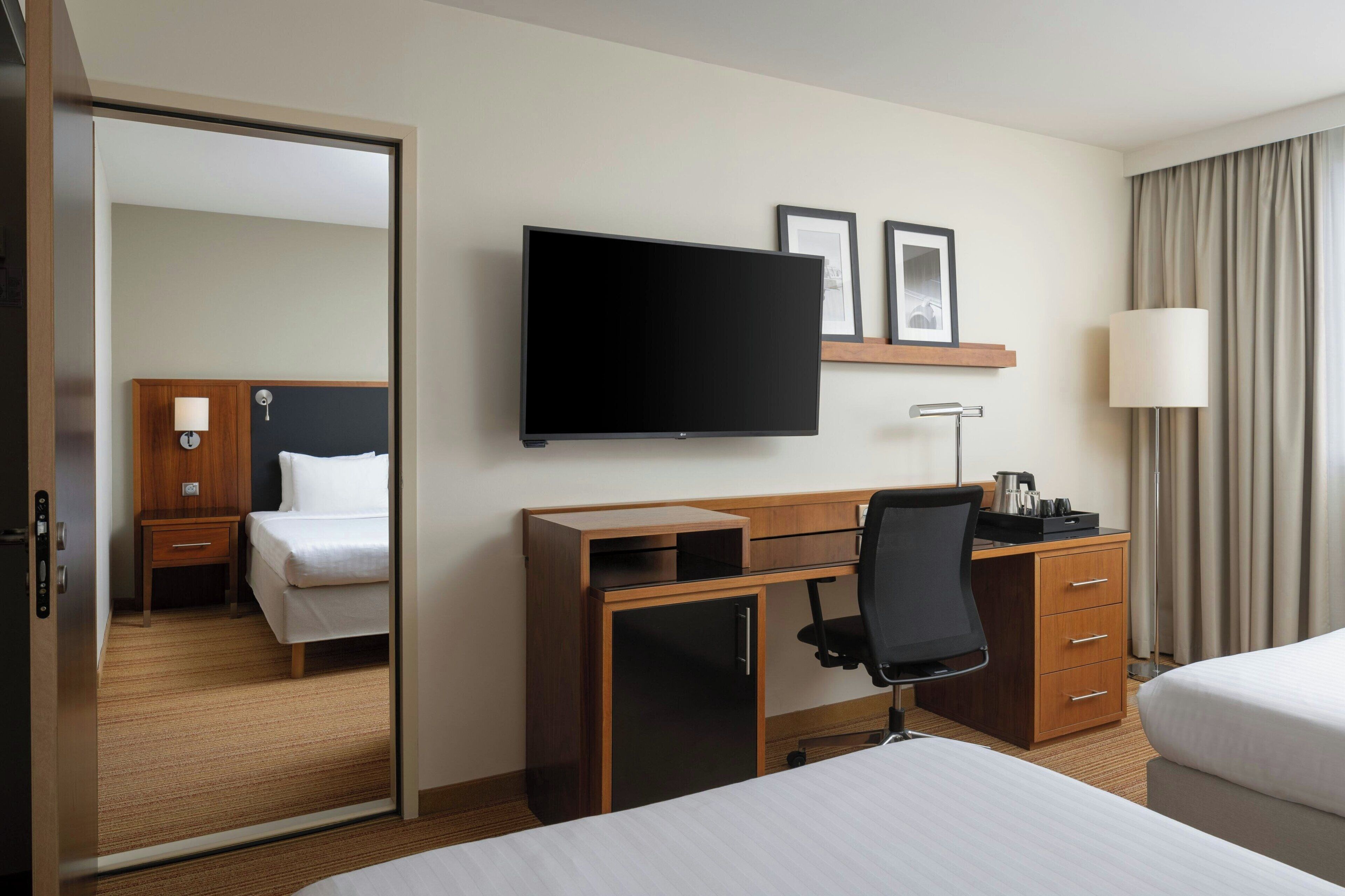 A hotel room with a bed, a desk with a chair, a television, a wardrobe, a bedside table, and a mirror.