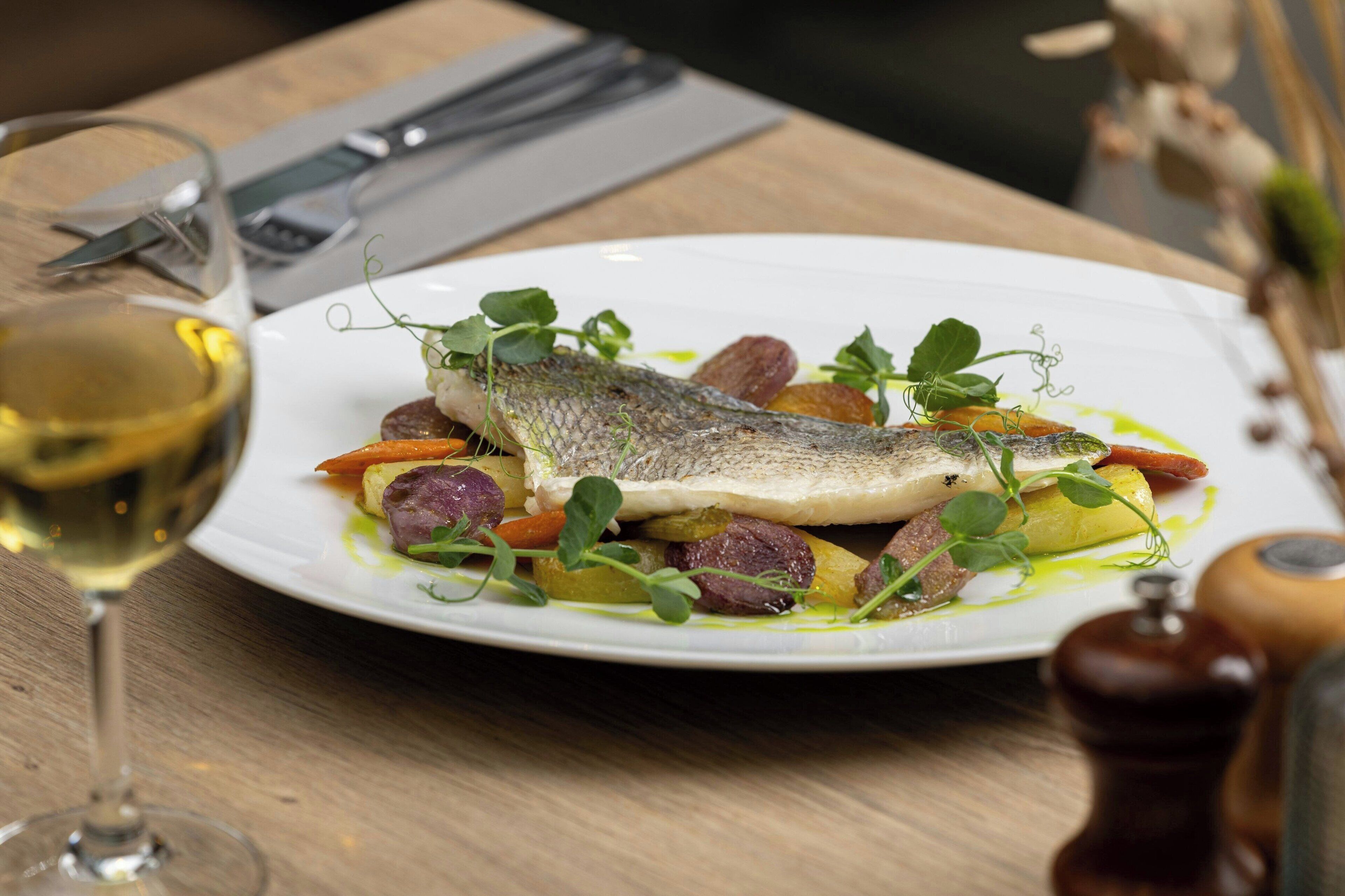 A plate of food with a fish, vegetables, and a glass of white wine on a wooden table.