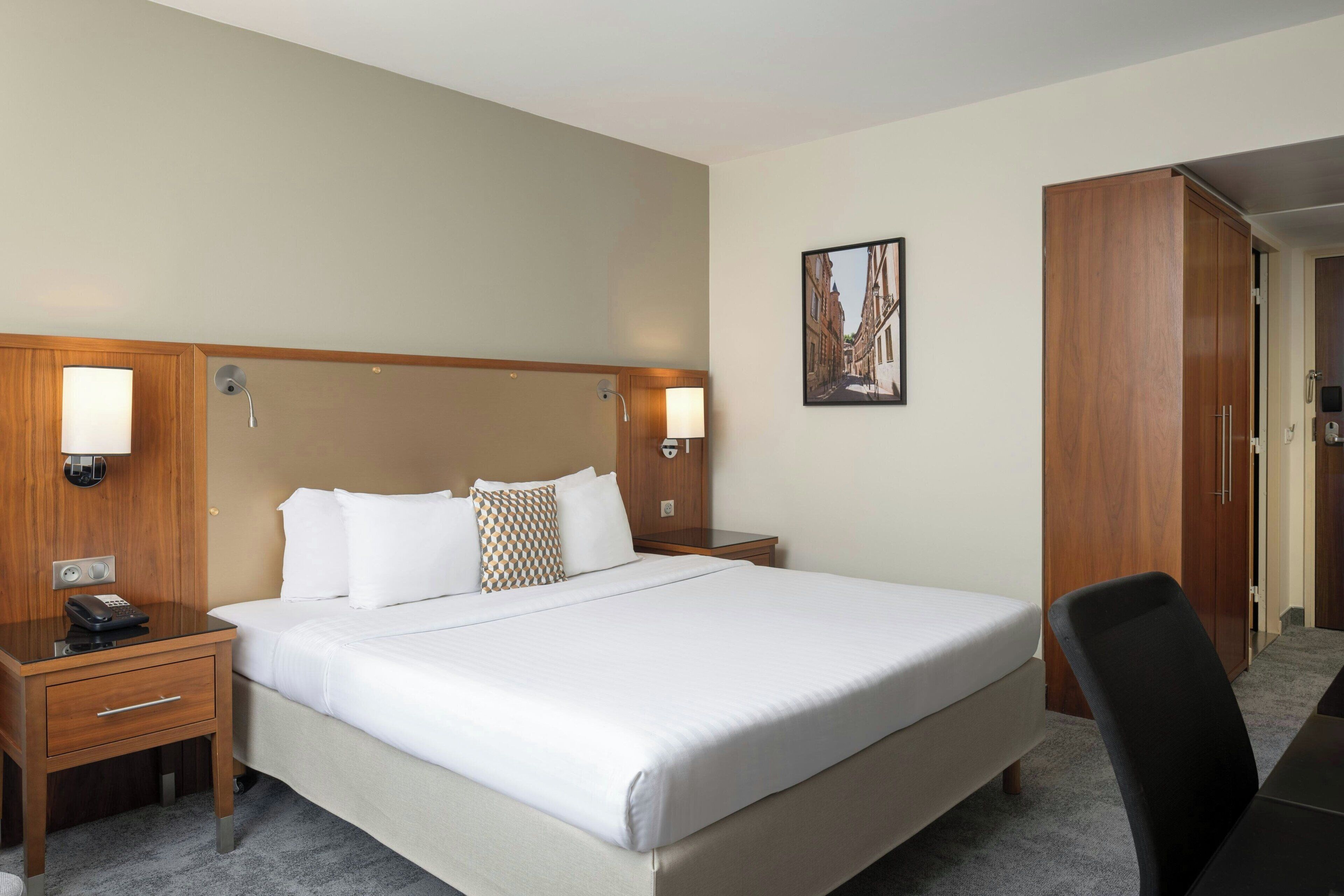 Una habitación de hotel con una cama grande, cabecera de madera, lámparas de noche, un escritorio con teléfono y un armario.