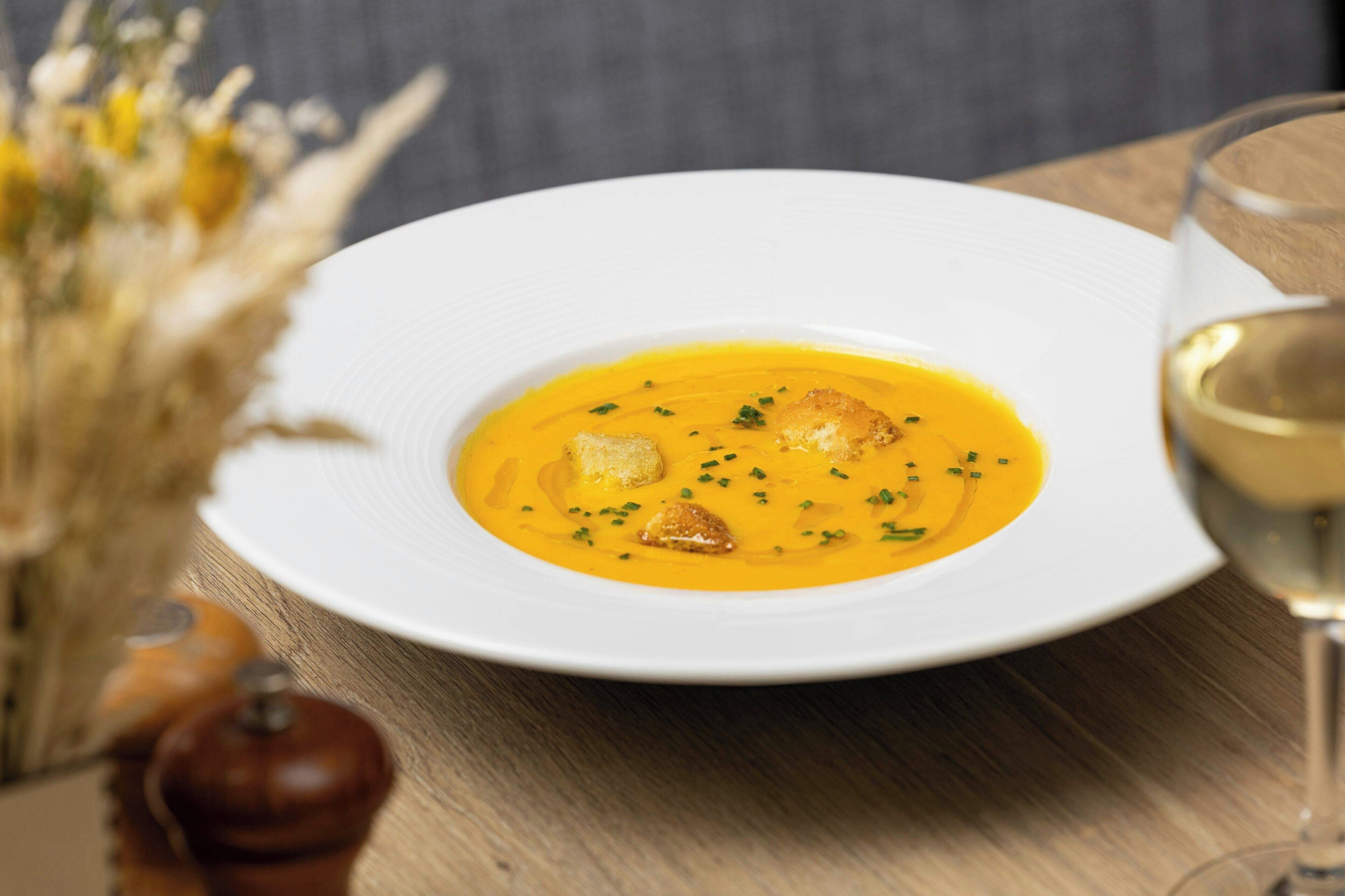 Un plato blanco con un tazón de sopa, una copa de vino blanco y un molinillo de pimienta sobre una mesa de madera.