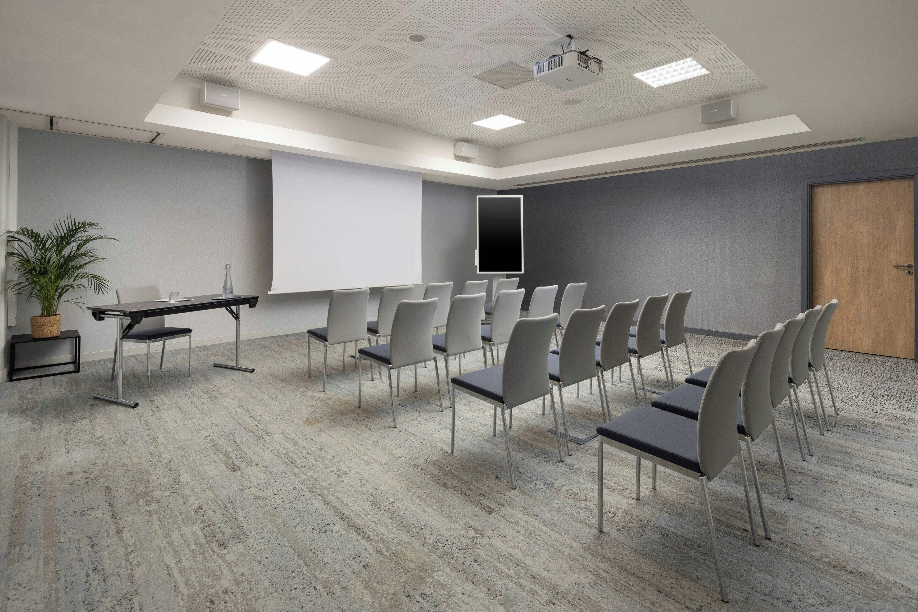 Une salle de conférence équipée d’un écran de projection, d’un tableau blanc et de rangées de chaises.