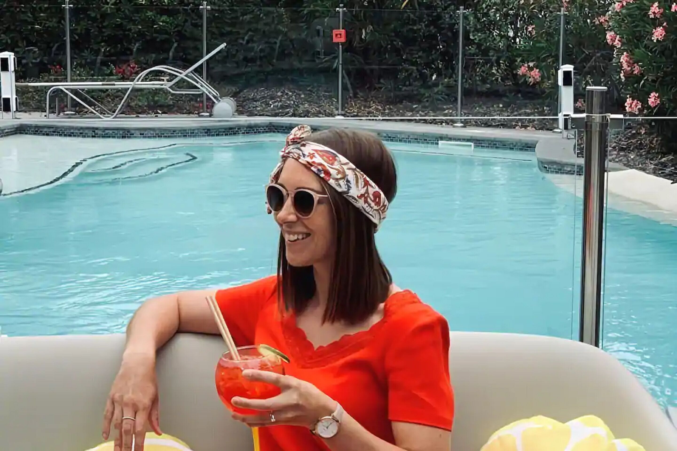 Una persona sentada junto a una piscina, disfrutando de una bebida, con un top rojo y una diadema.