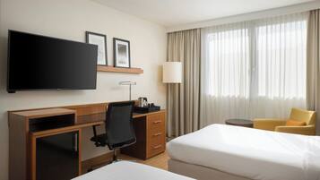 Quarto de hotel com cama, escrivaninha, cadeira, televisão, abajur e janela com cortinas.