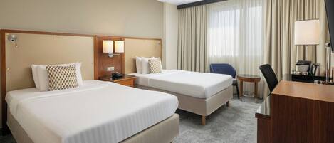 A hotel room with two beds, a desk, a chair, a small table, and a TV.