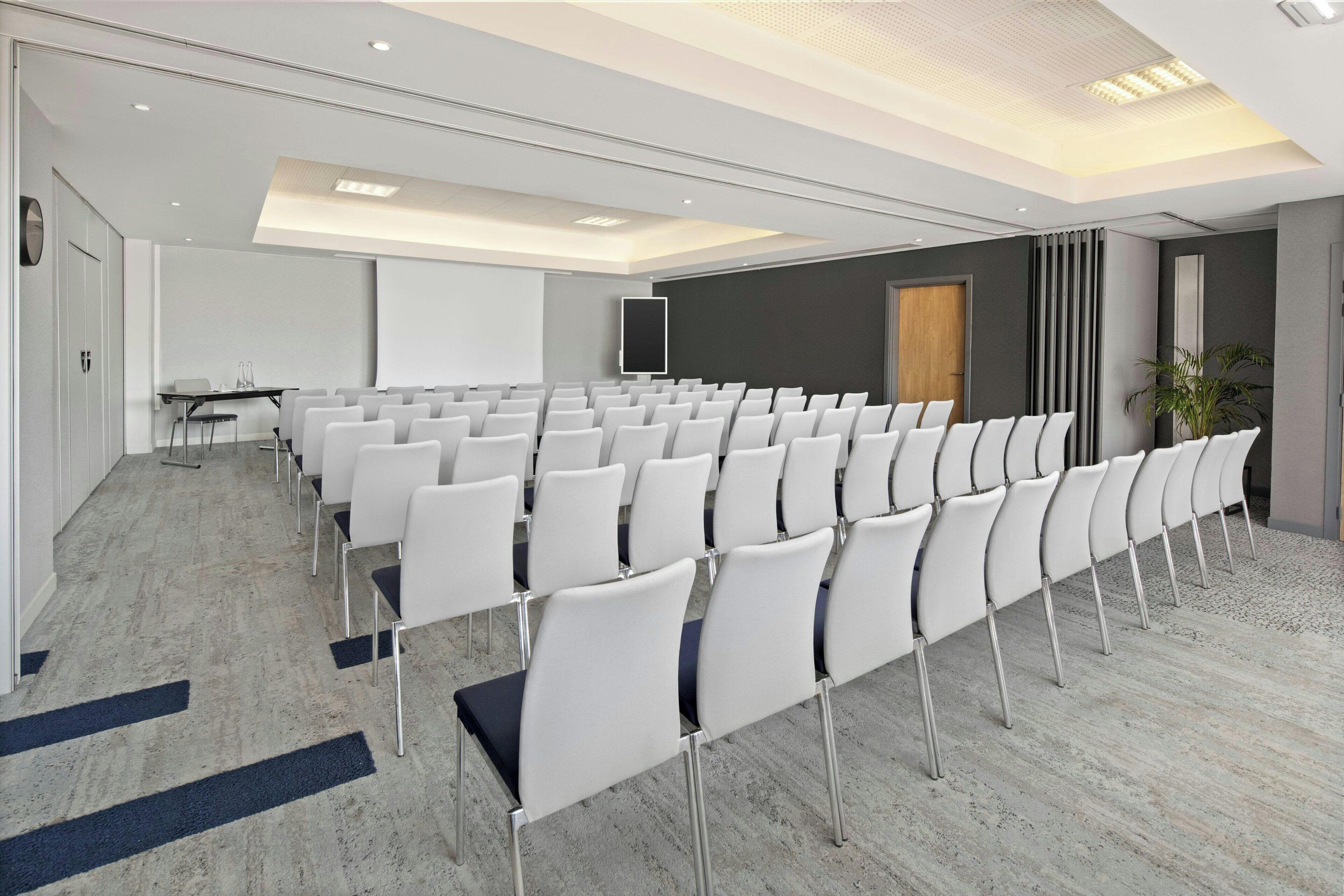A conference room with rows of white chairs, a projector screen, and a table with chairs.