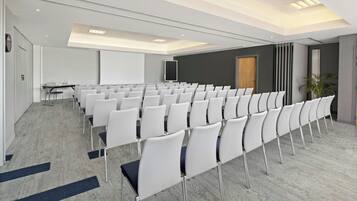 A conference room with rows of white chairs, a projector screen, and a table with chairs.