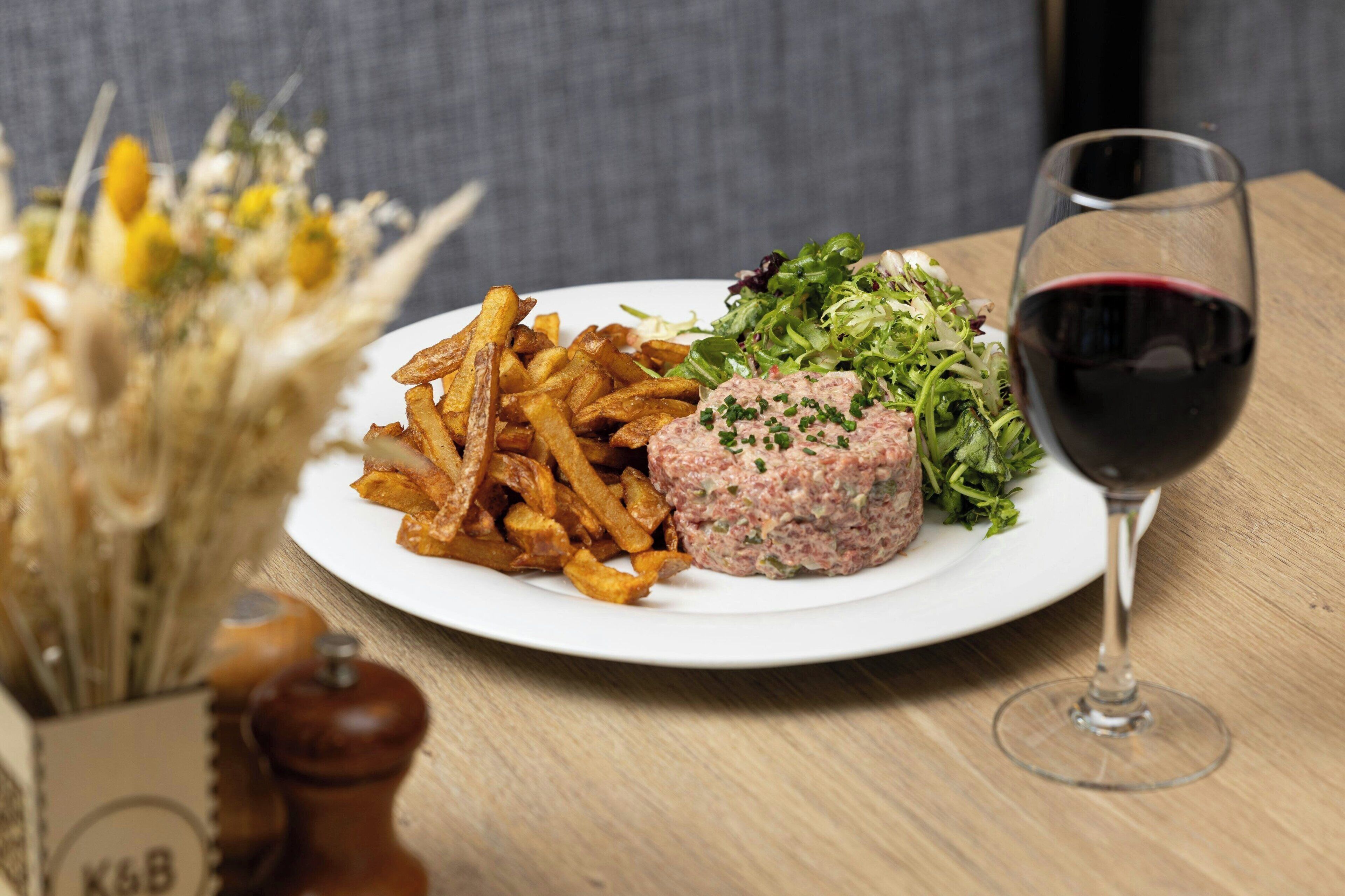 Un plato de comida con papas fritas, una hamburguesa y ensalada, acompañado de una copa de vino tinto sobre una mesa de madera.