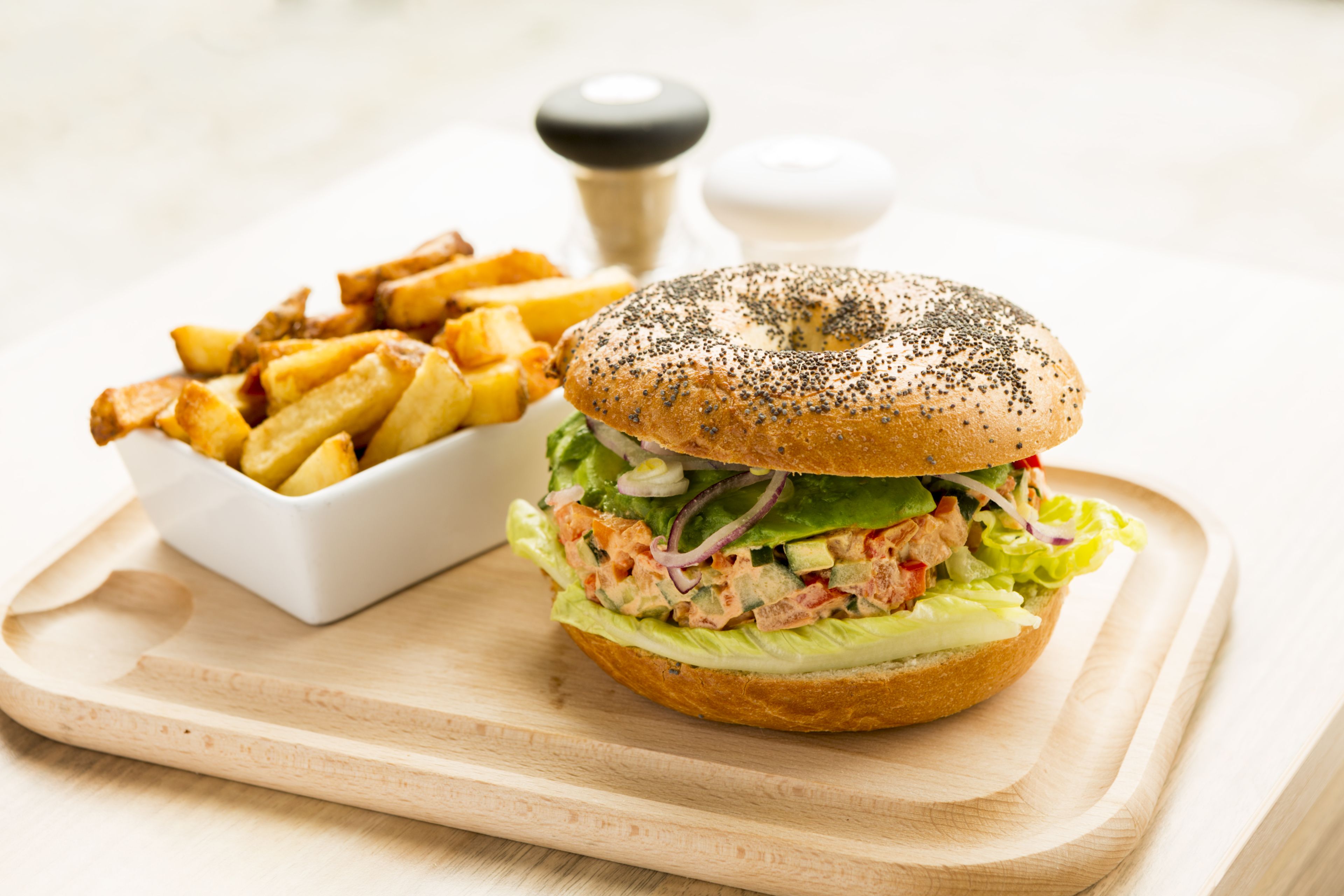 A sandwich with lettuce, tomato, and a poppy seed bun, alongside a bowl of fries on a wooden board.
