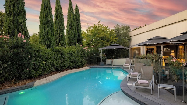 A swimming pool with lounge chairs and umbrellas, surrounded by tall trees and a building with a modern design.