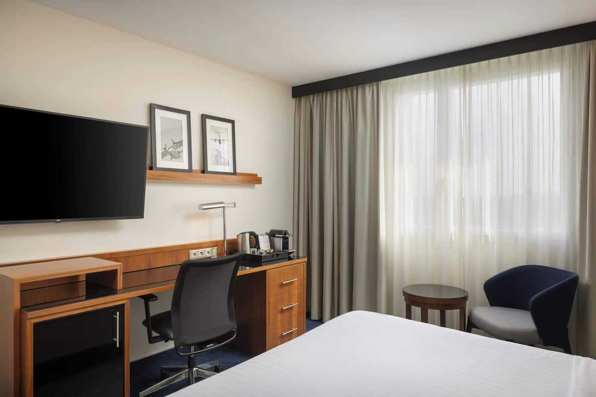 Une chambre d’hôtel comprenant un lit, un bureau avec un ordinateur, une chaise, une télévision et une fenêtre avec des rideaux.