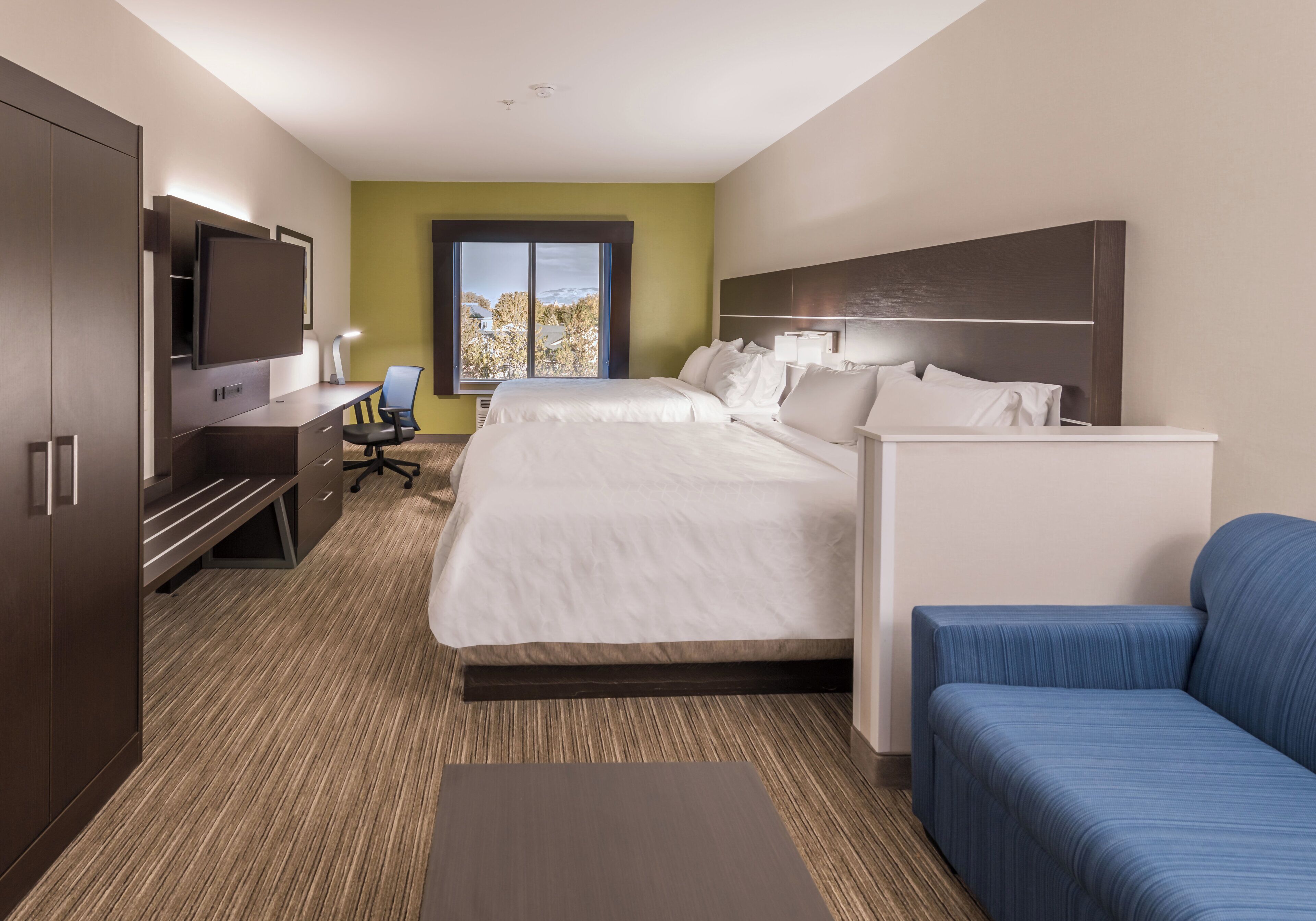 A hotel room with a bed, a desk with a chair, a TV, and a window with a view of trees.