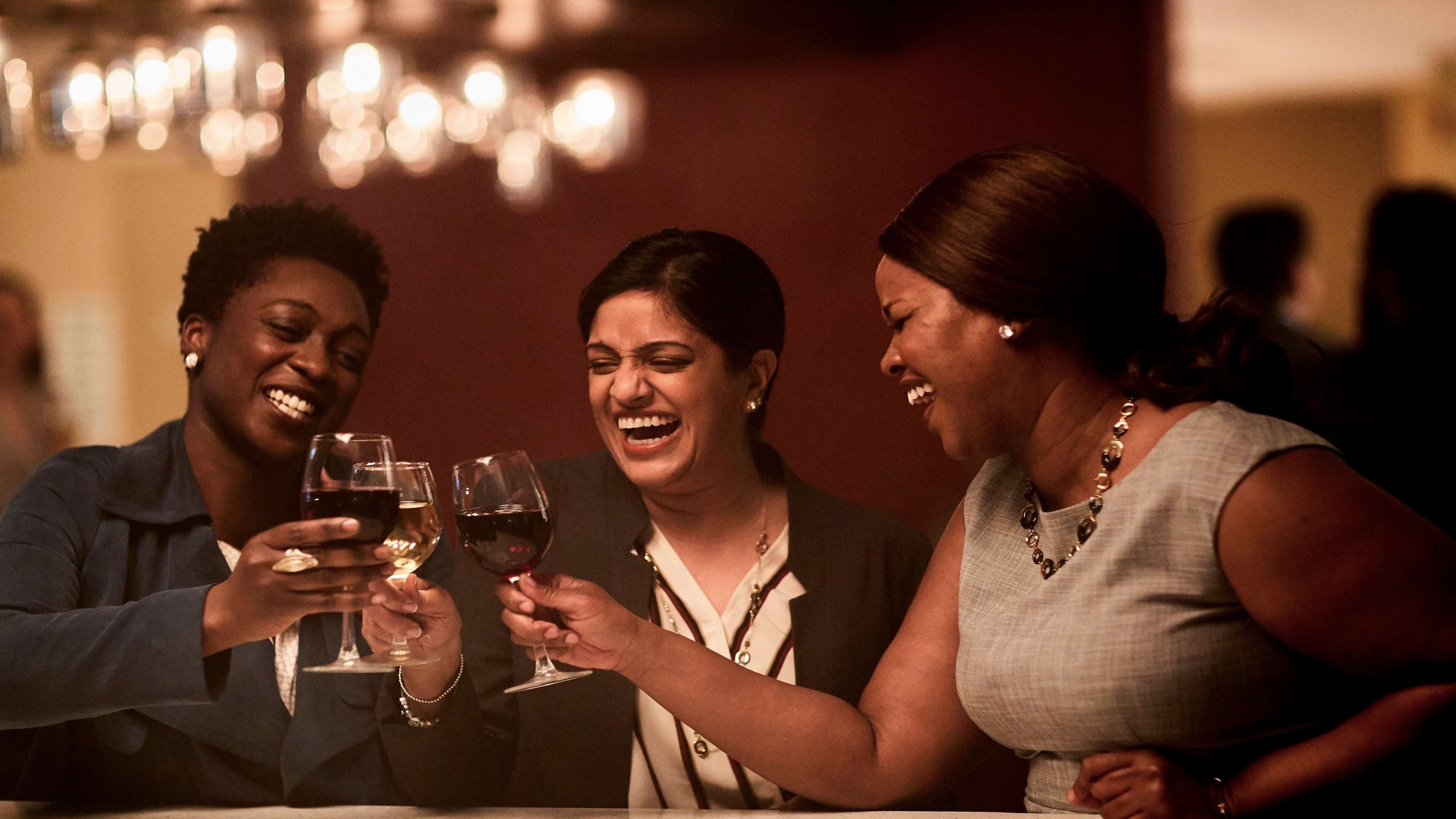 Trois personnes trinquent avec des verres à vin autour d’une table dans une pièce faiblement éclairée, où un lustre est suspendu au plafond.