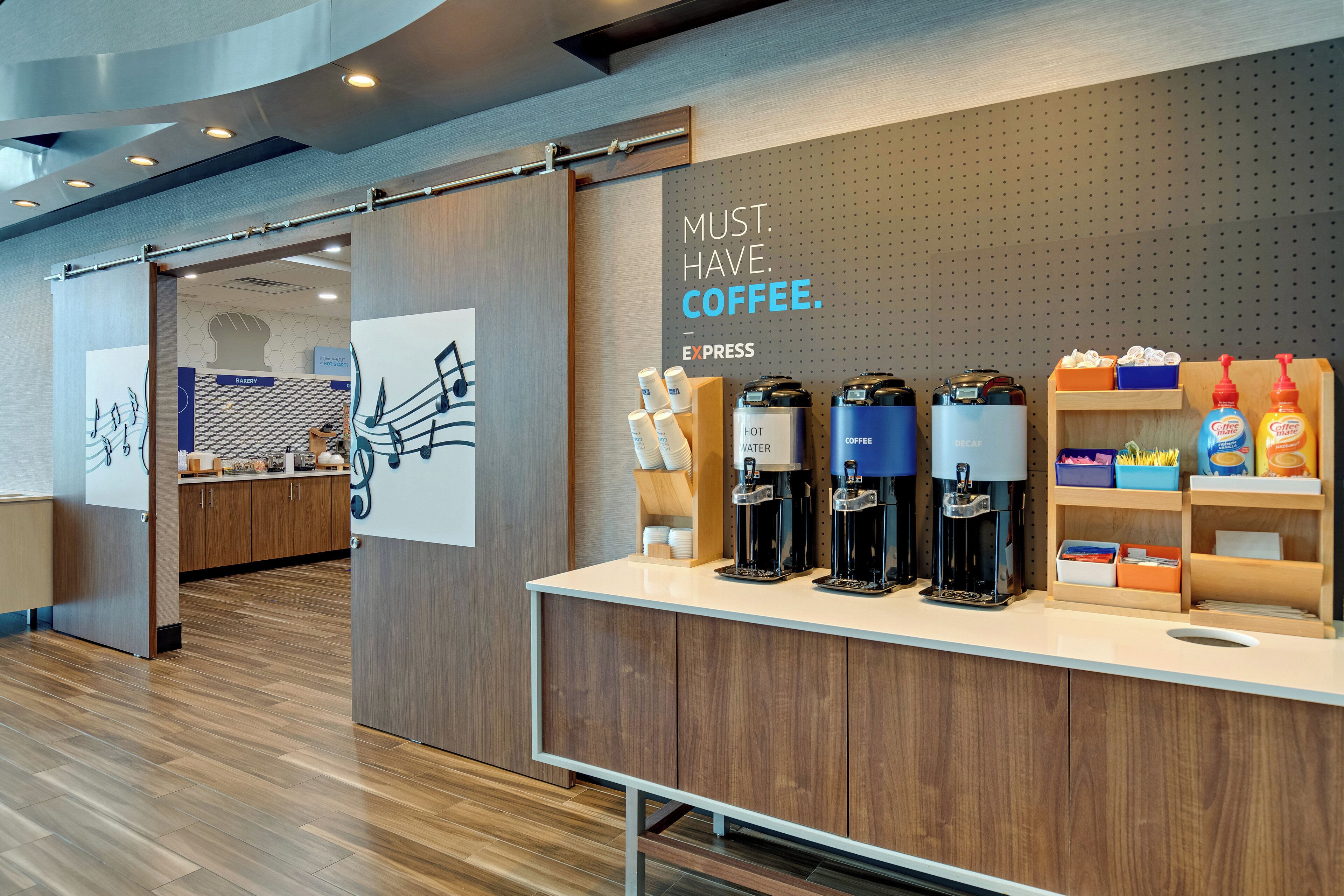 Una estación de café con múltiples dispensadores, un cartel que dice 'MOST. HAVE. COFFEE.' y una pared con diseño hexagonal.