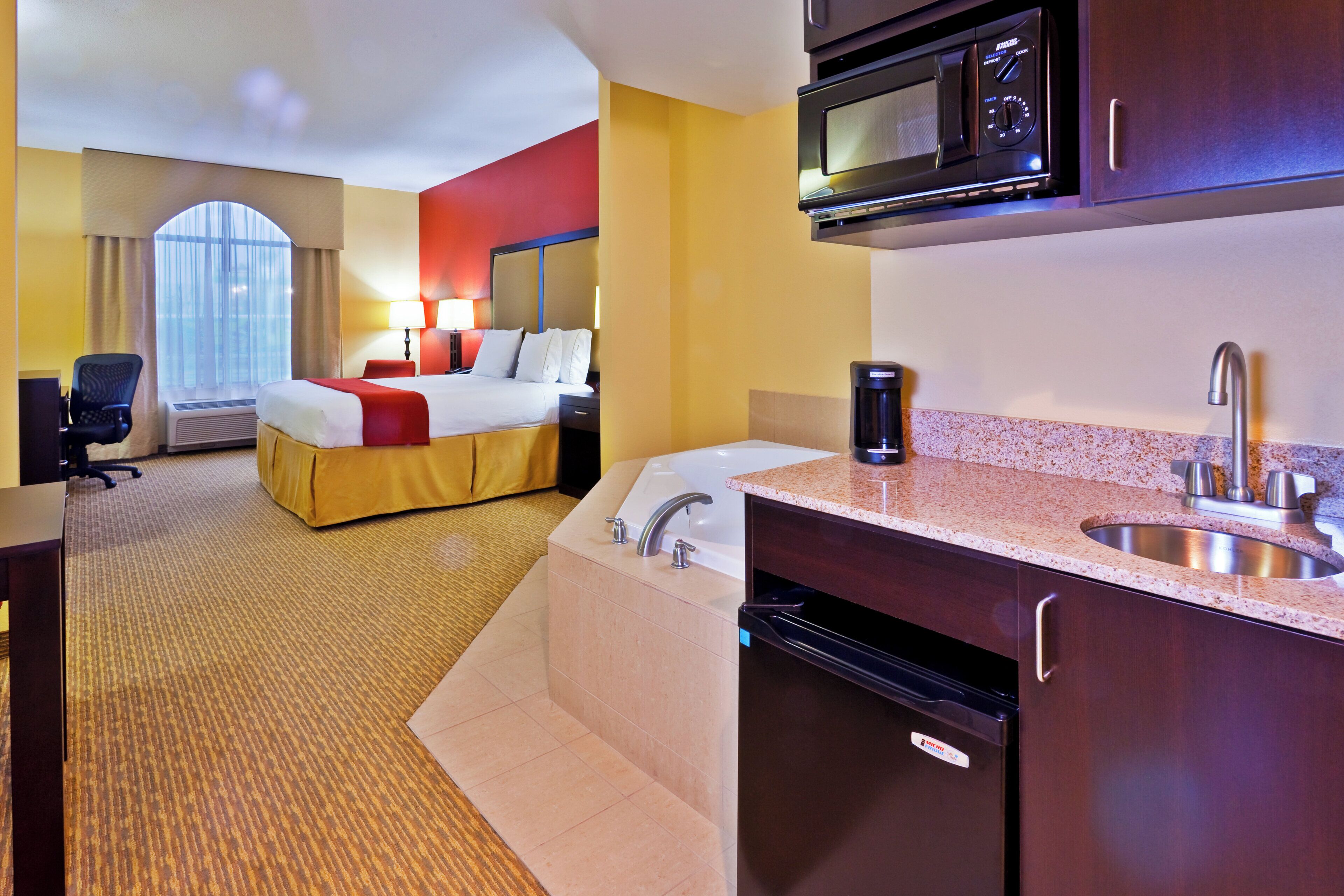 A hotel room with a bed, a desk, a microwave, a sink, and a view of the outdoors.