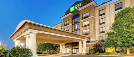 Ein Holiday Inn Express Gebäude mit überdachter Eingangspartie und einer begrünten Anlage mit Bäumen und Sträuchern.