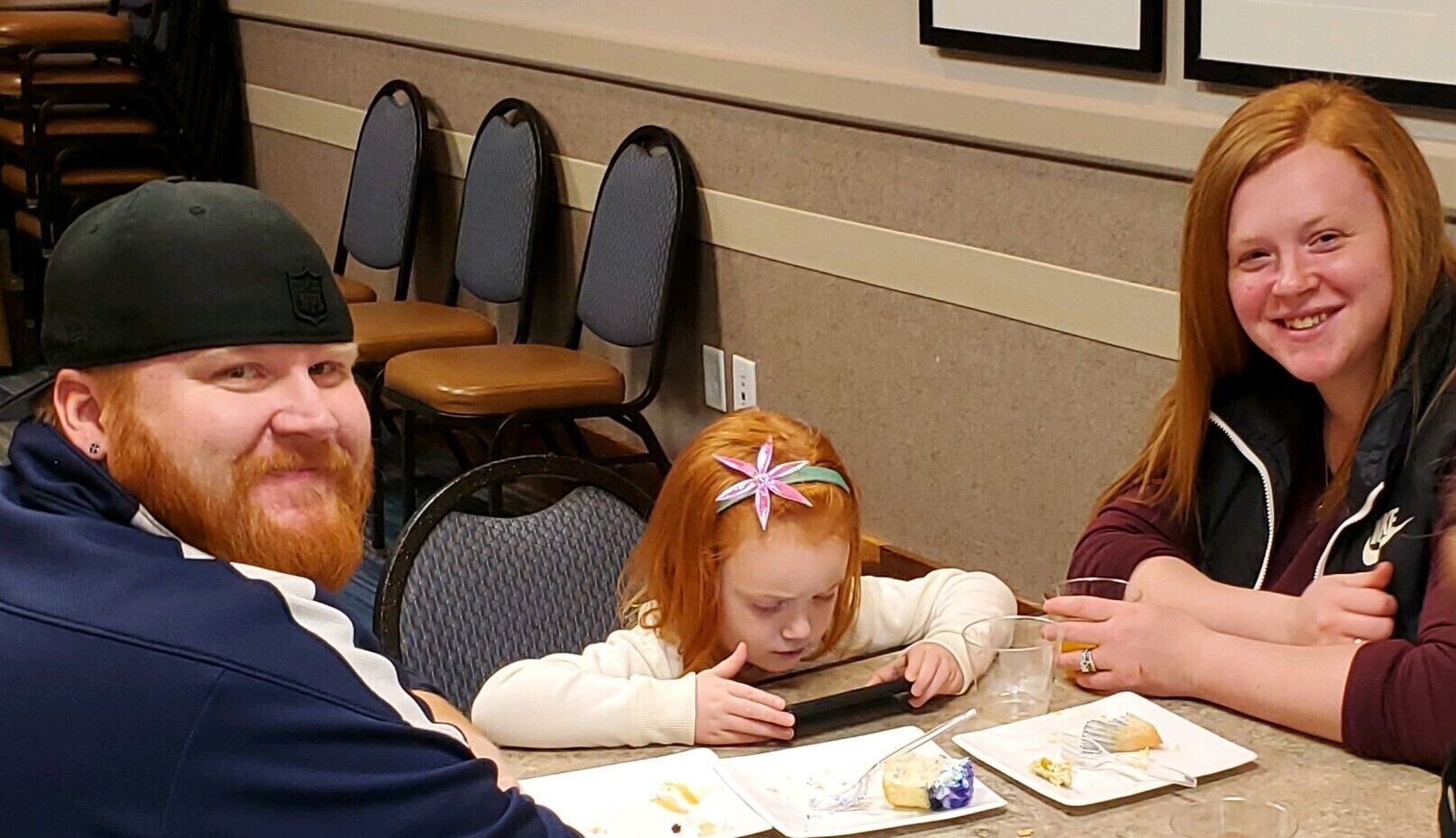 Tres personas sentadas a una mesa con platos de comida, sillas y un enchufe en la pared.