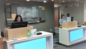 A reception desk with a computer, a bottle of hand sanitizer, and a framed picture. There is a wall with a bulletin board and a sign above the desk. - Holiday Inn Express Hotel and Suites Mankato East by IHG (Mankato)