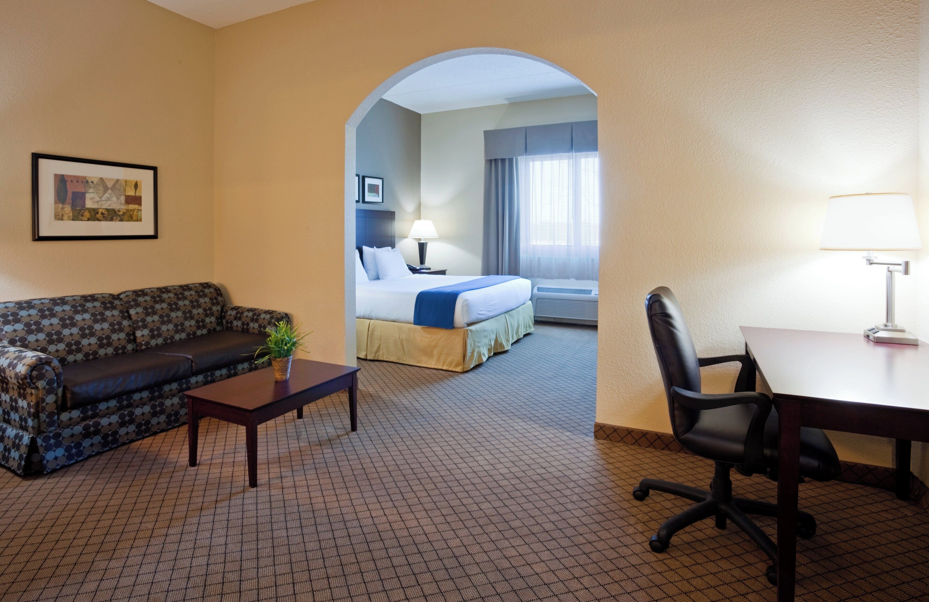A hotel room with a sofa, a desk, a bed, and an arched doorway leading to another room.