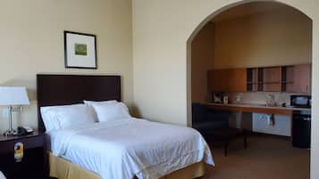 Quarto de hotel com cama, mesa de cabeceira, luminária, quadro na parede e um arco que dá para uma cozinha americana com micro-ondas e pia.