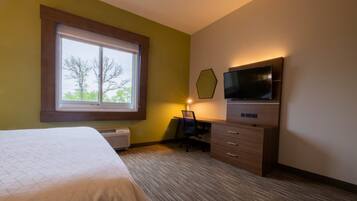 A hotel room with a bed, a desk with a chair, a TV, and a window with a view of trees.