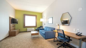 A hotel room with a blue sofa, a wooden desk, a computer, a TV, and a window with natural light.