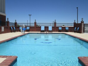 Una piscina rectangular con agua azul cristalina, rodeada por un muro de ladrillo y una valla metálica. Hay sillones de descanso y sombrillas a lo largo de la piscina.