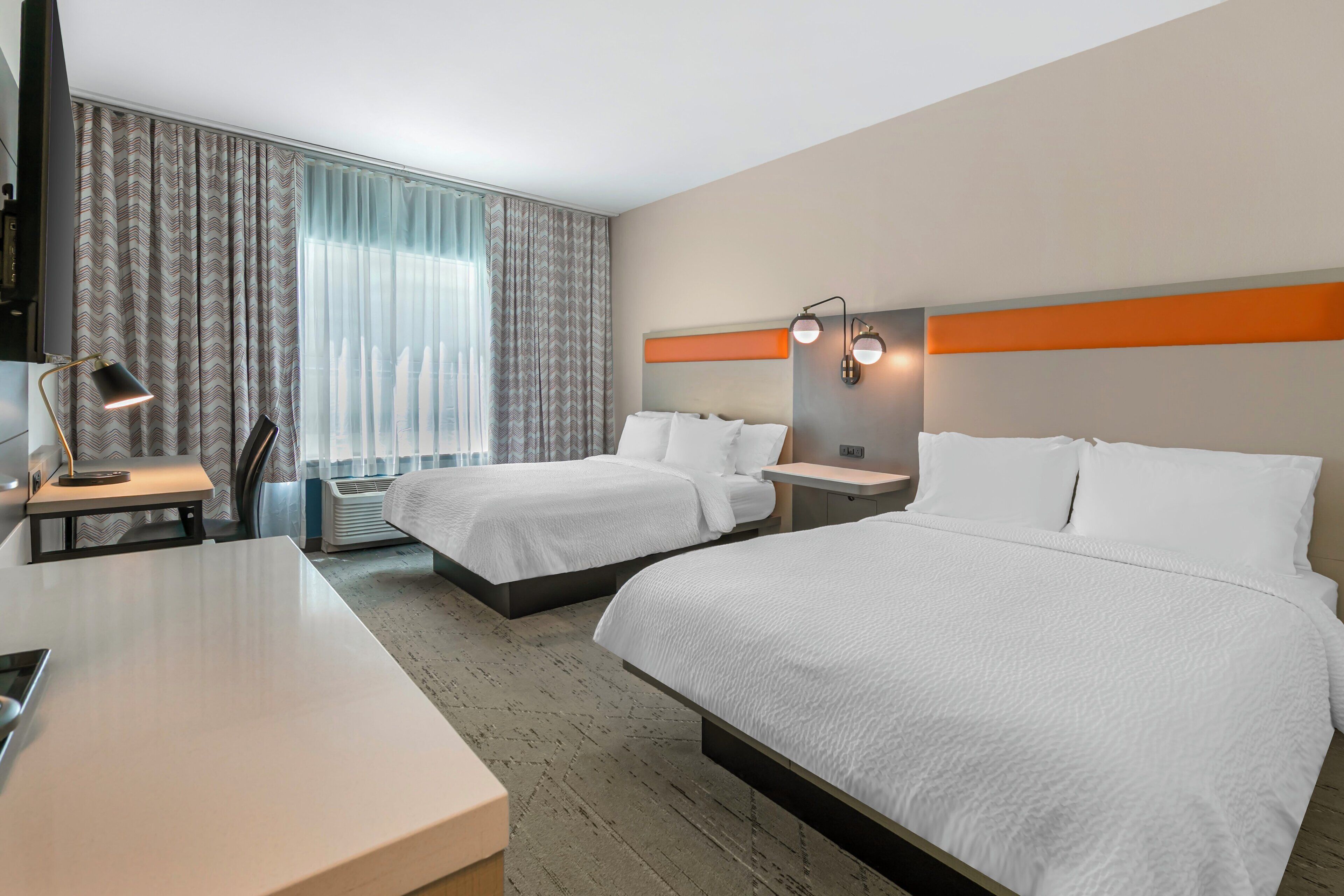 A hotel room with two beds, a desk, a chair, a TV, and a window with curtains.