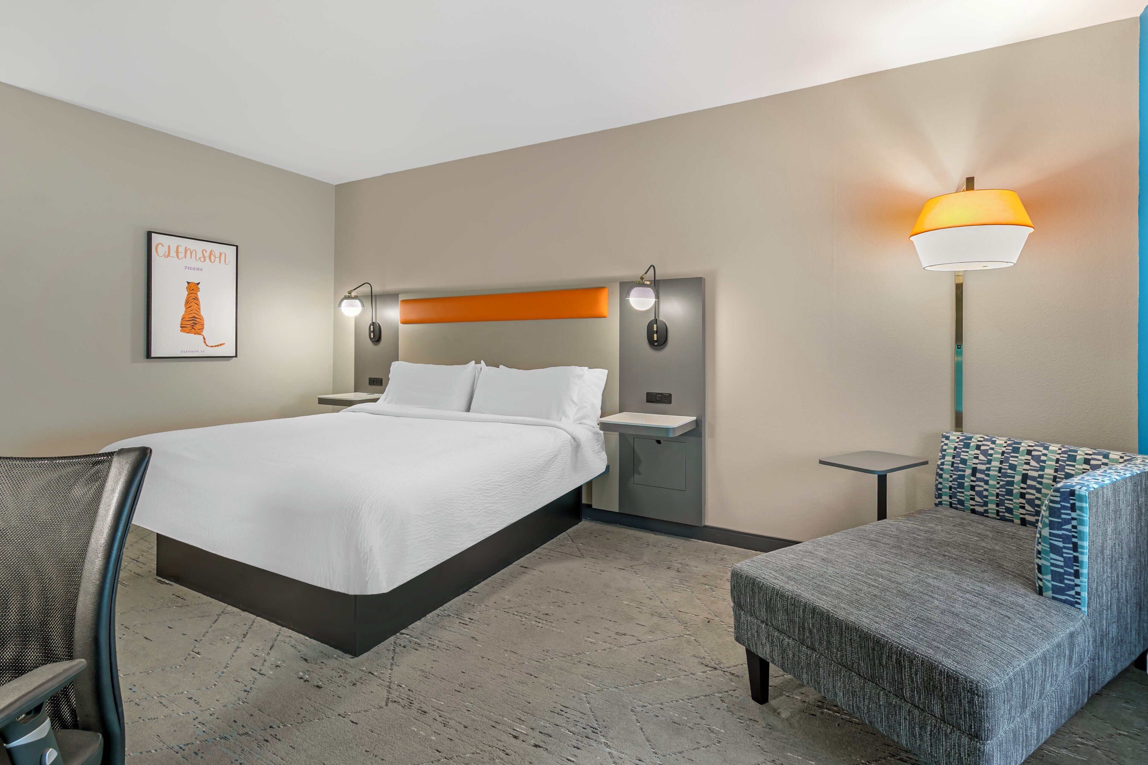 A hotel room with a bed, bedside tables, a chair, a small table, and a wall-mounted lamp.