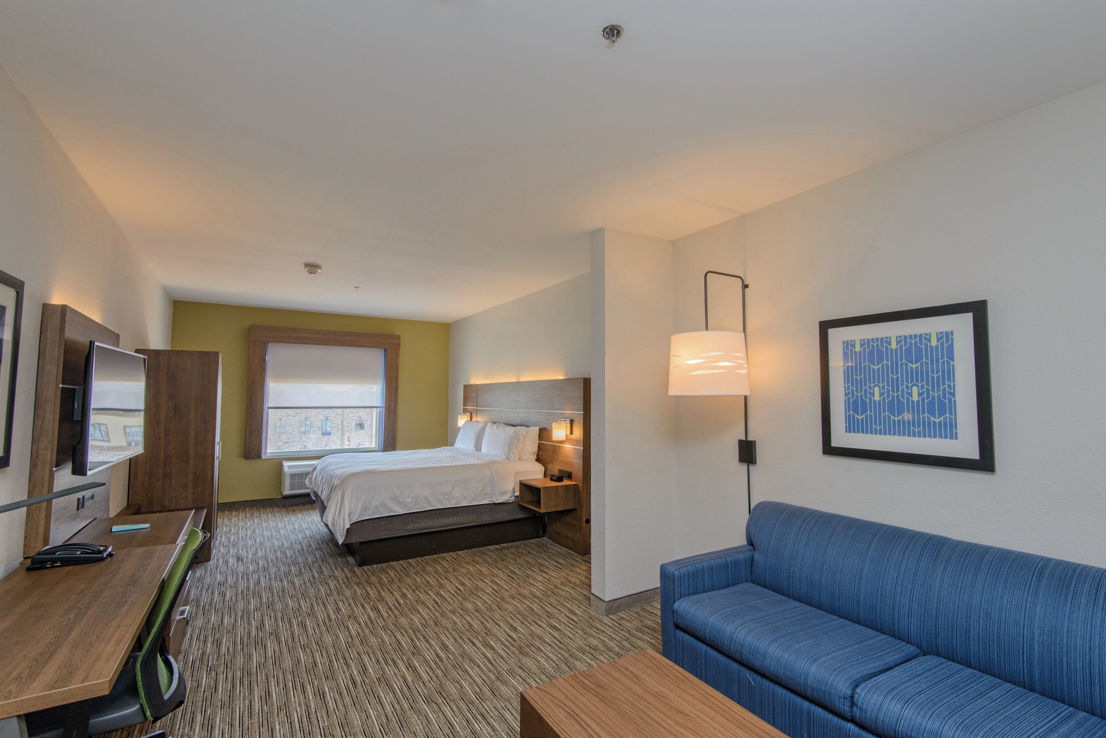 A hotel room with a bed, a sofa, a desk with a computer, a TV, and a framed picture on the wall.