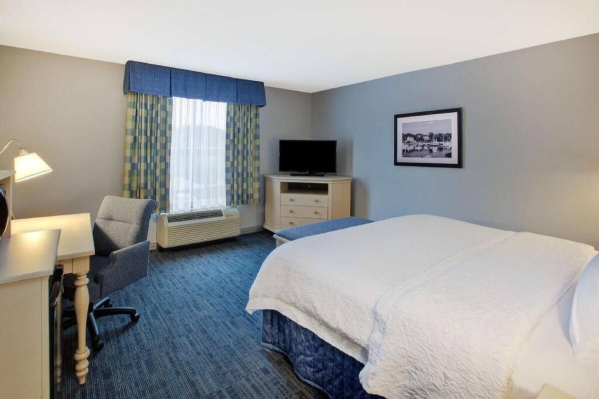 Une chambre d’hôtel avec un lit, un bureau doté d’une lampe, une chaise, un téléviseur posé sur un meuble et un tableau encadré accroché au mur.