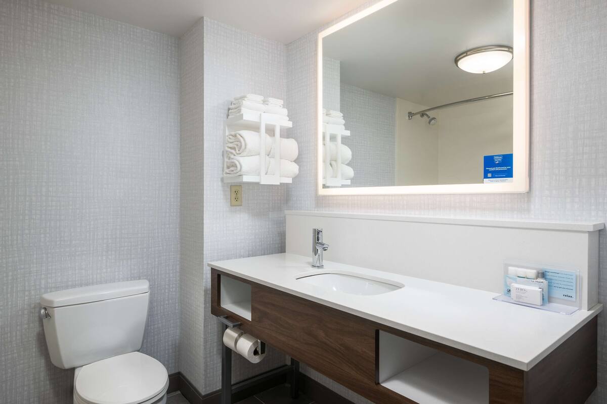 Une salle de bain avec des toilettes blanches, un lavabo surmonté d’un miroir et un porte-serviettes avec des serviettes.