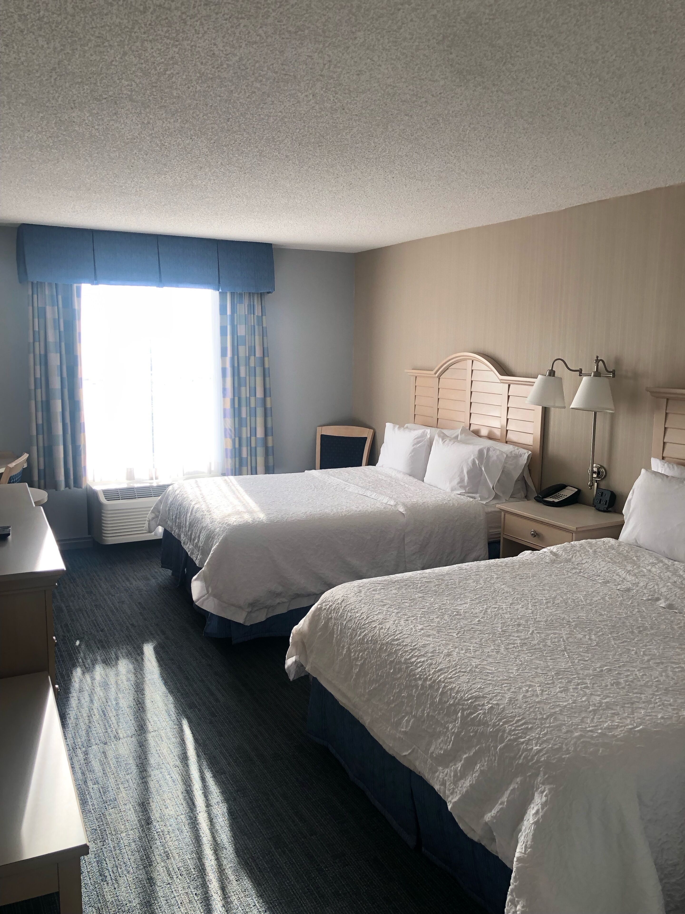A hotel room with two beds, a desk, a chair, a nightstand with a lamp, and a window with curtains.