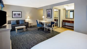 A hotel room with a bed, a desk, a chair, a sofa, a TV, and a bathroom with a mirror and sink.