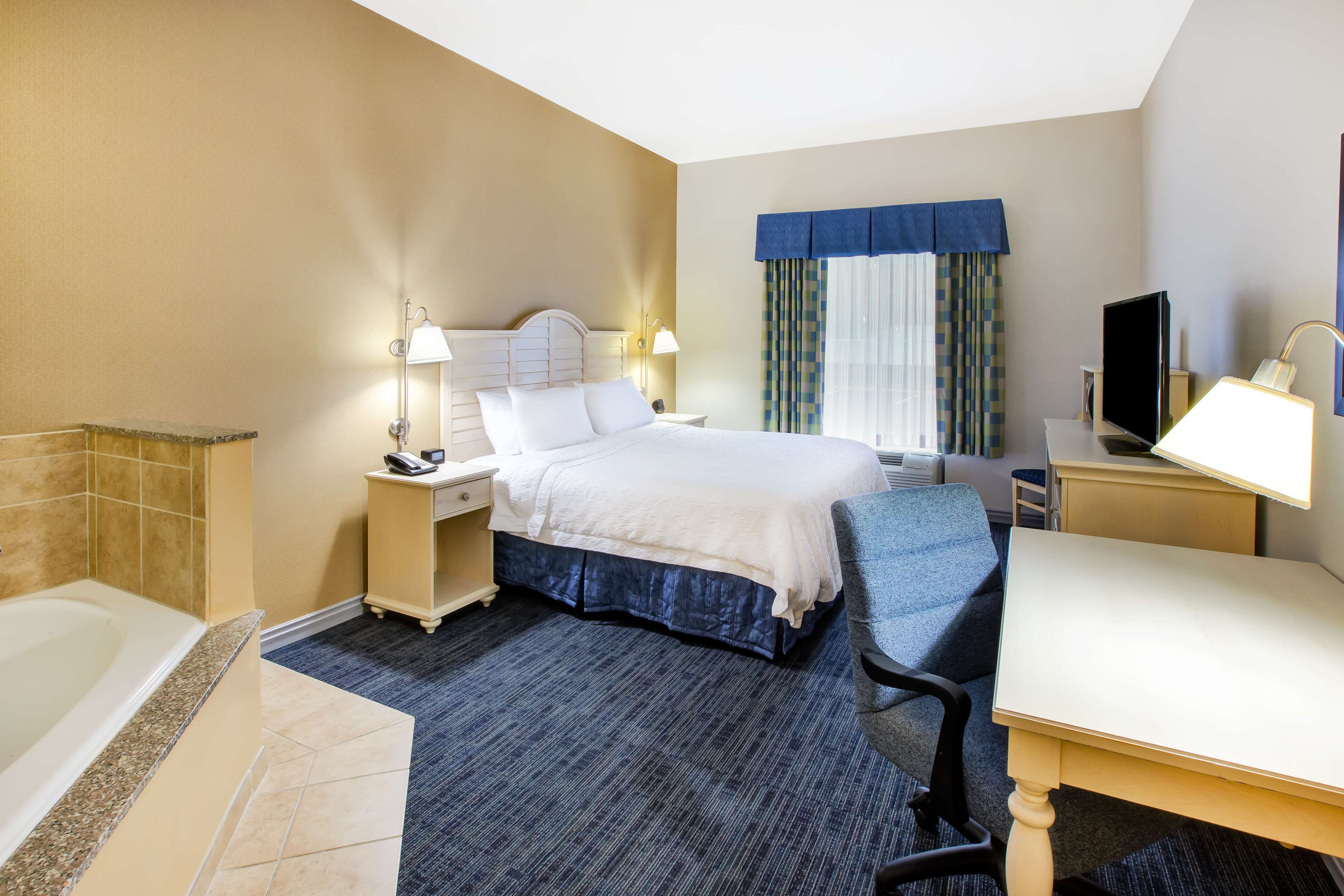 A hotel room with a bed, a desk, a chair, a TV, a lamp, a nightstand, and a bathroom with a bathtub.