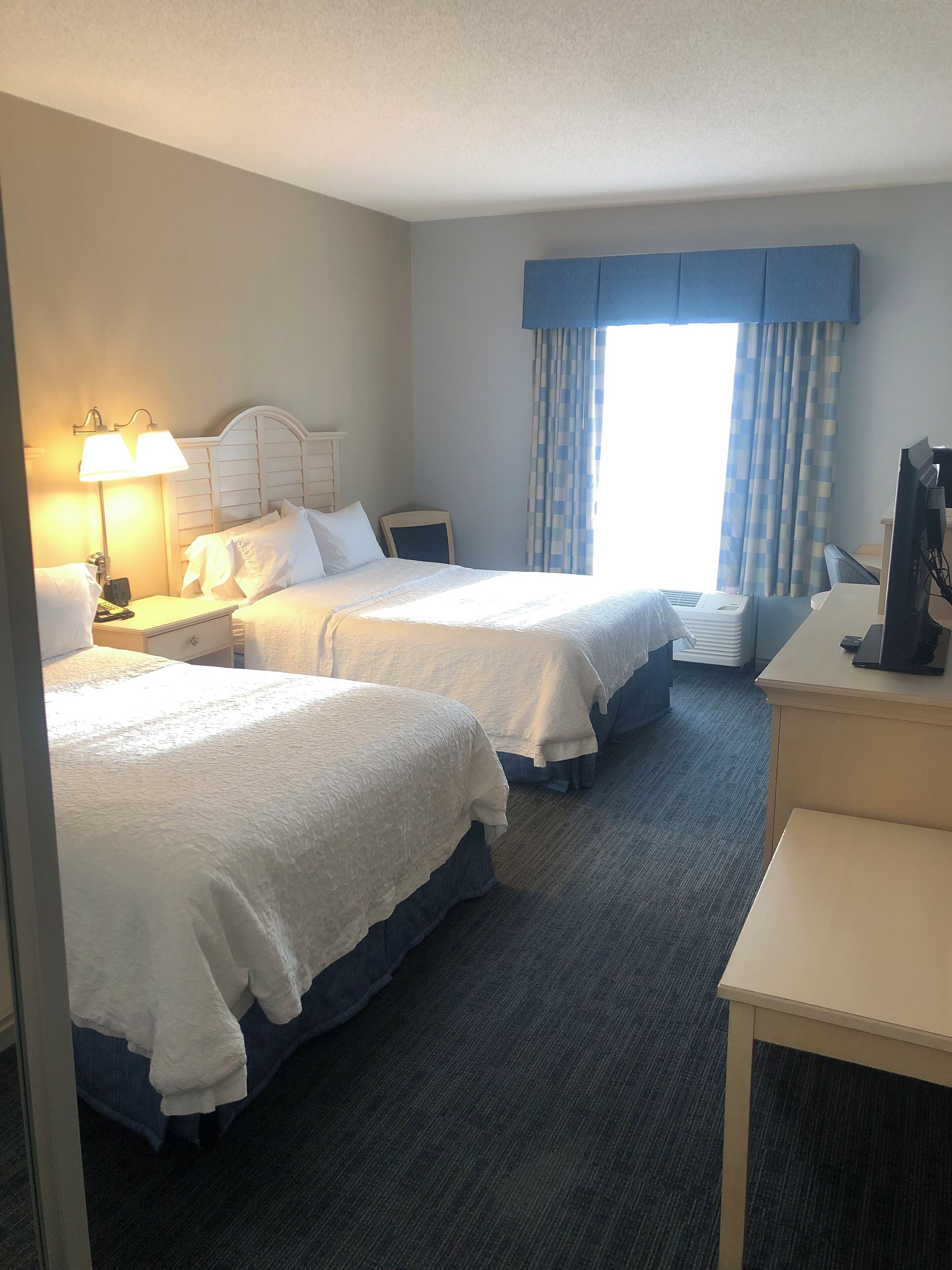 A hotel room with two beds, a desk, a chair, a lamp, and a window with curtains.