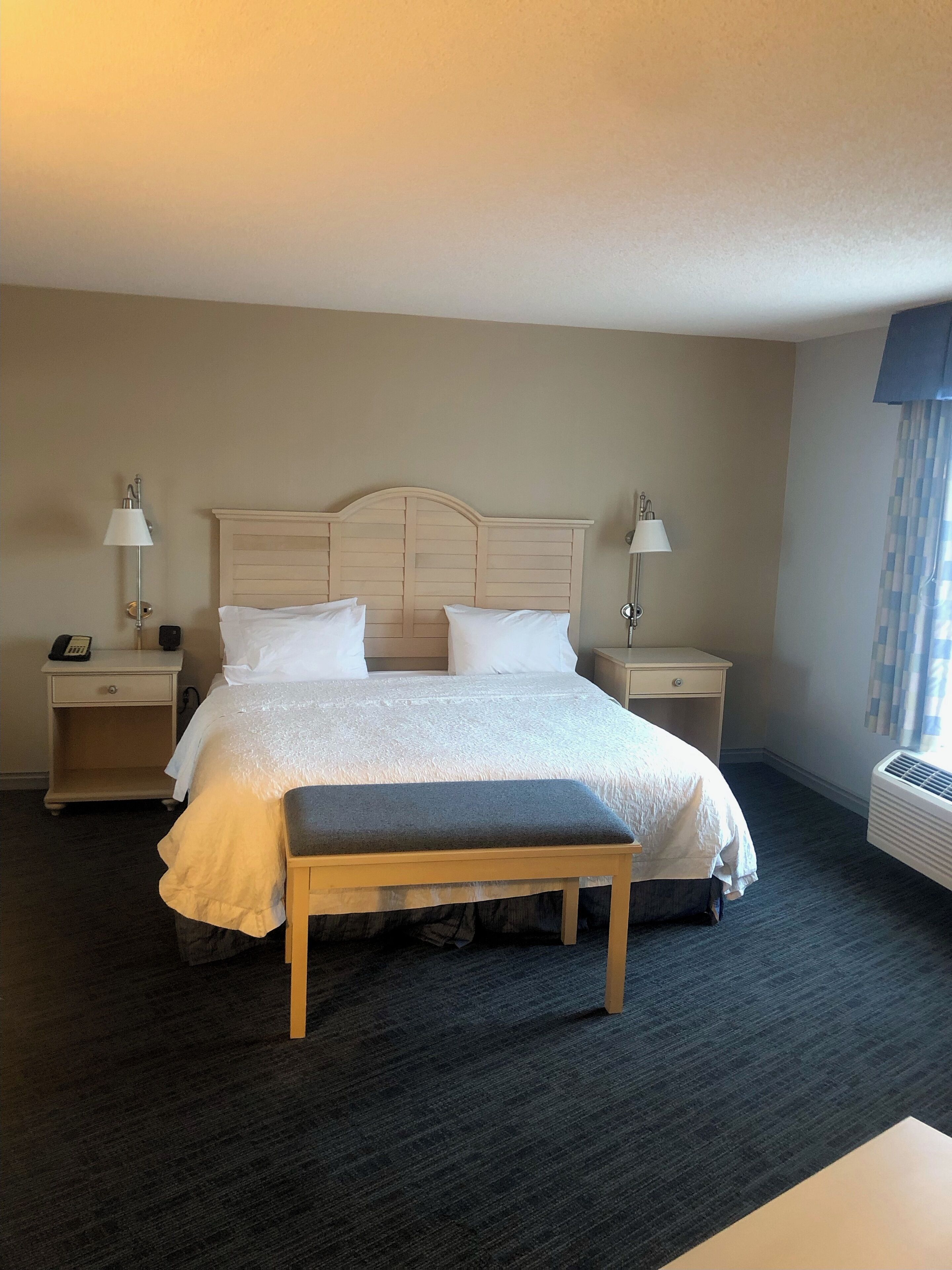 Habitación de hotel con una cama grande, mesitas de noche con lámparas, un banco y un equipo de aire acondicionado.