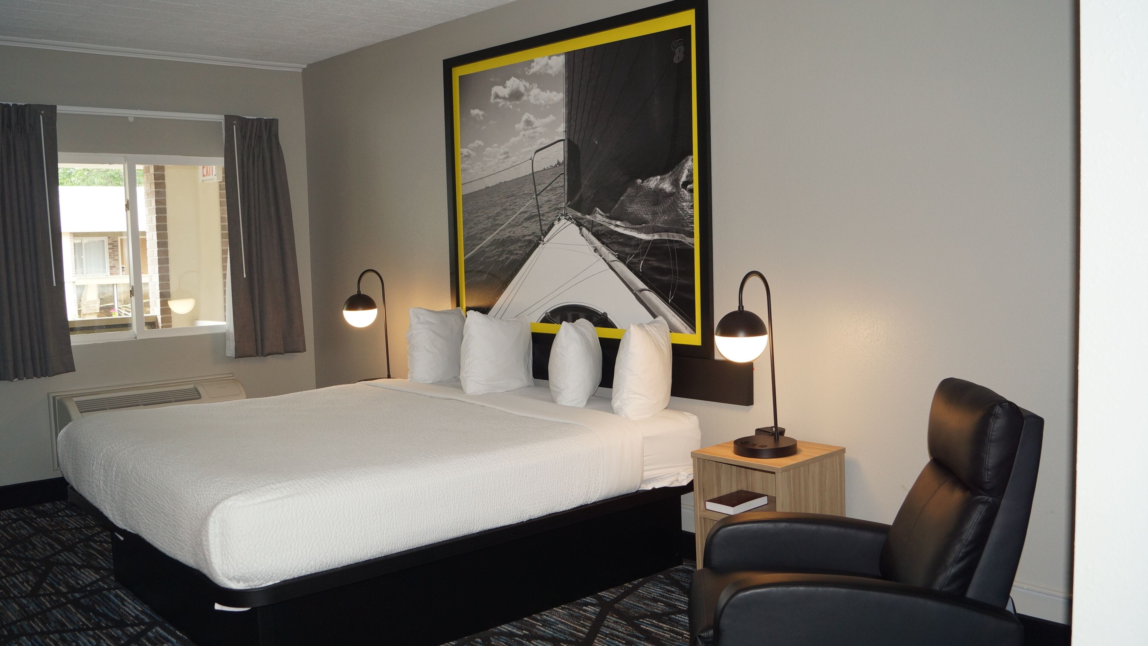 A hotel room with a large bed, two bedside lamps, a chair, a small table, and a large framed picture on the wall.