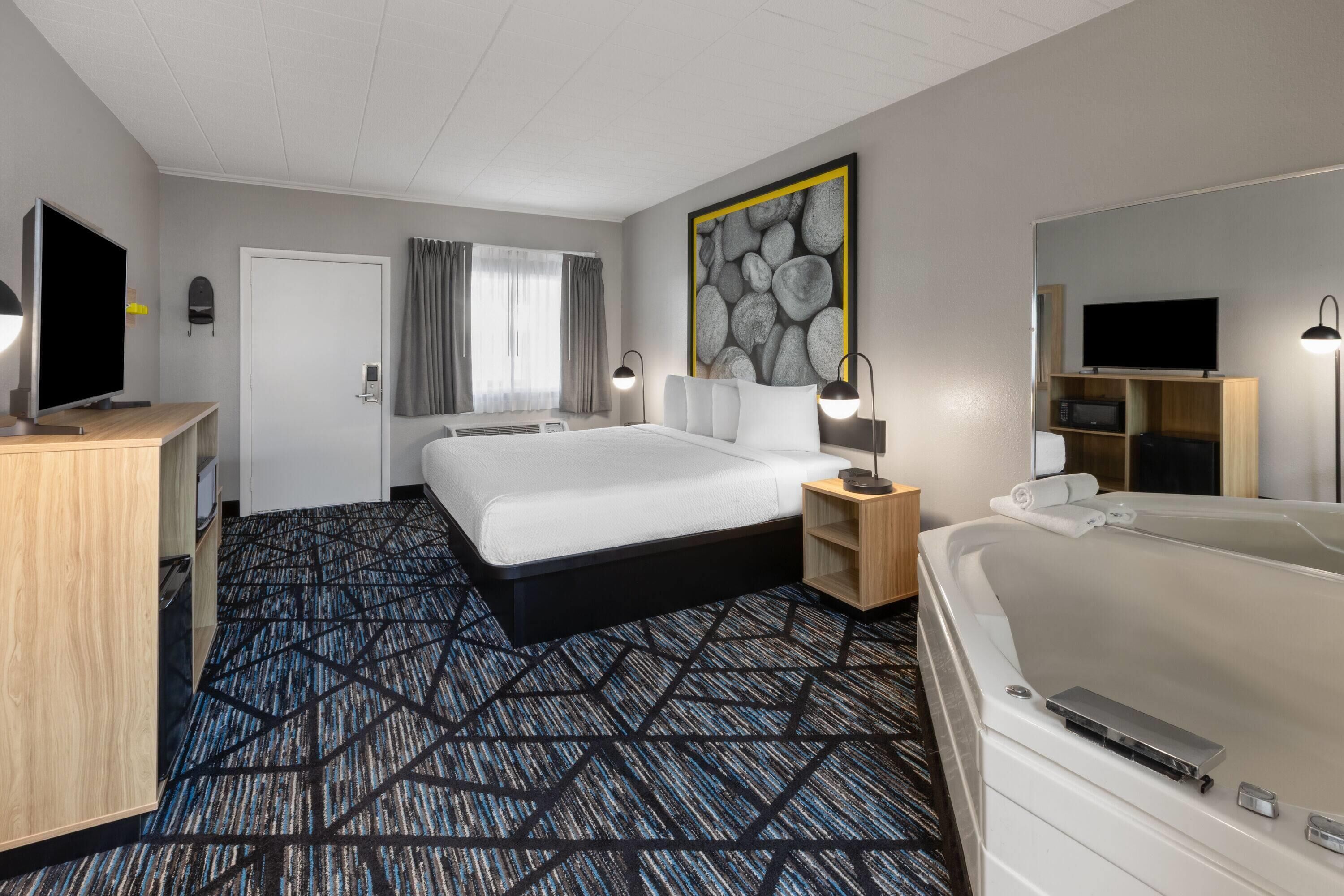 A hotel room with a large bed, a desk with a flat-screen TV, a desk lamp, a wall-mounted TV, a bathtub, and a large artwork on the wall.