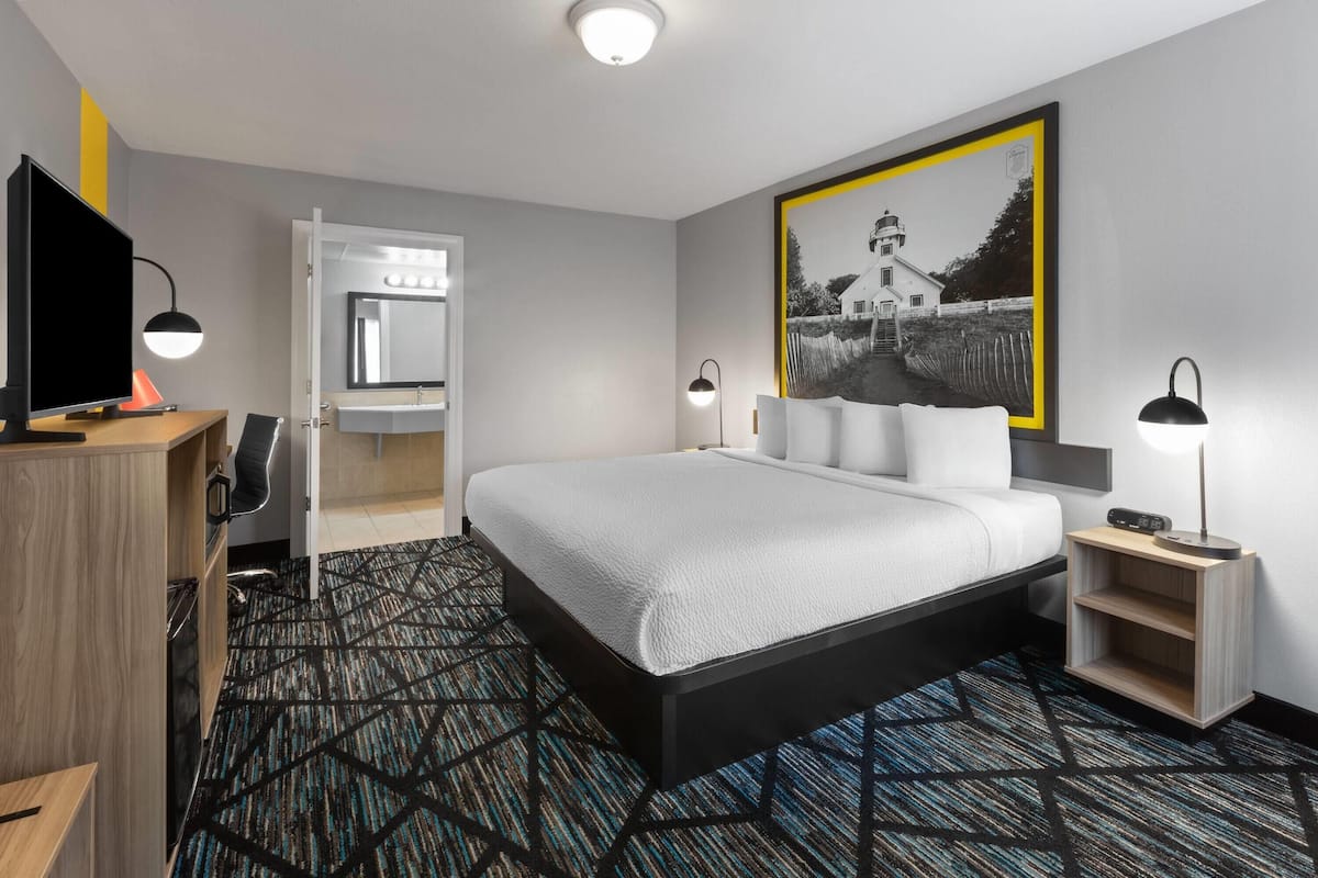 A modern hotel room with a large bed, a desk with a computer, a wardrobe, and a bathroom mirror.