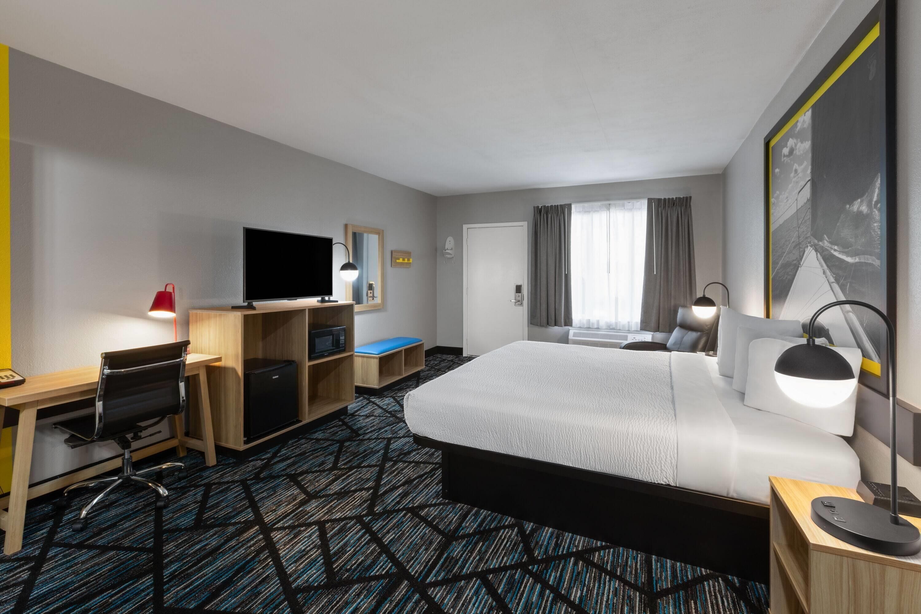 Habitación de hotel con una cama grande, un escritorio con computadora, un televisor, una lámpara de escritorio y una ventana con cortinas.