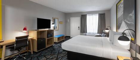 Habitación de hotel con una cama grande, un escritorio con computadora, un televisor, una lámpara de escritorio y una ventana con cortinas.