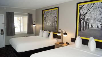 A hotel room with two beds, a window with curtains, a bedside table with a lamp, and two framed pictures on the wall.