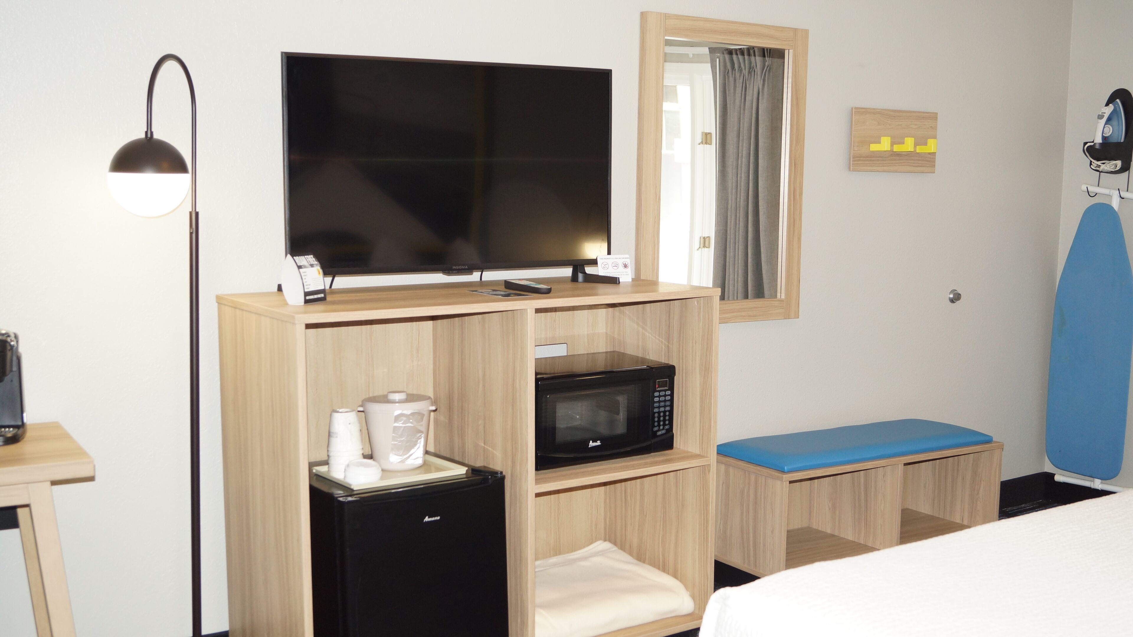 A compact hotel room with a flat-screen TV, a microwave, a mini-fridge, a small table with a coffee maker, a blue bench, and a surfboard.