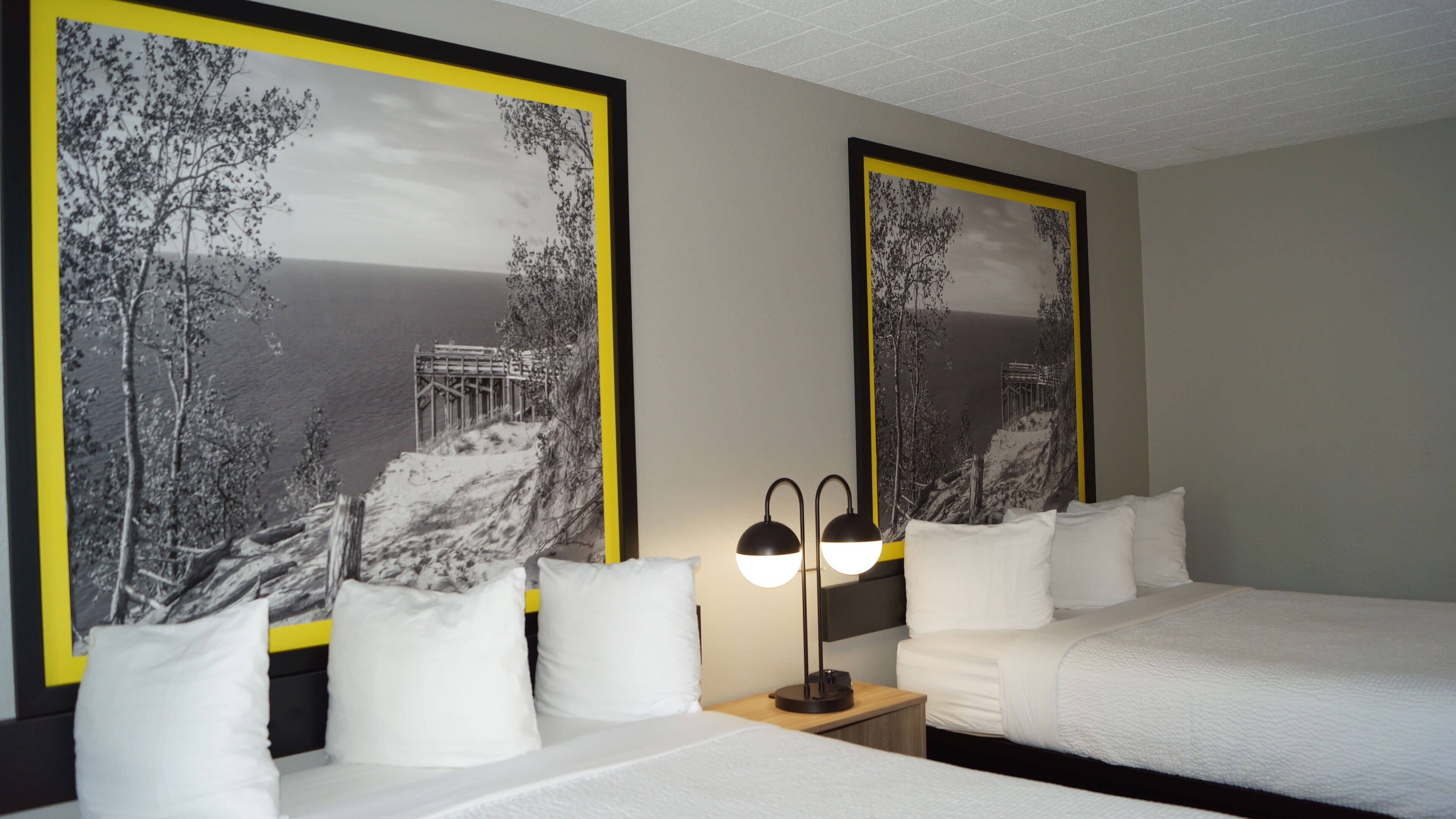 Une chambre d’hôtel avec deux lits, du linge de lit blanc, une table de chevet avec une lampe, et de grandes peintures encadrées en noir et blanc représentant des paysages, accrochées aux murs.
