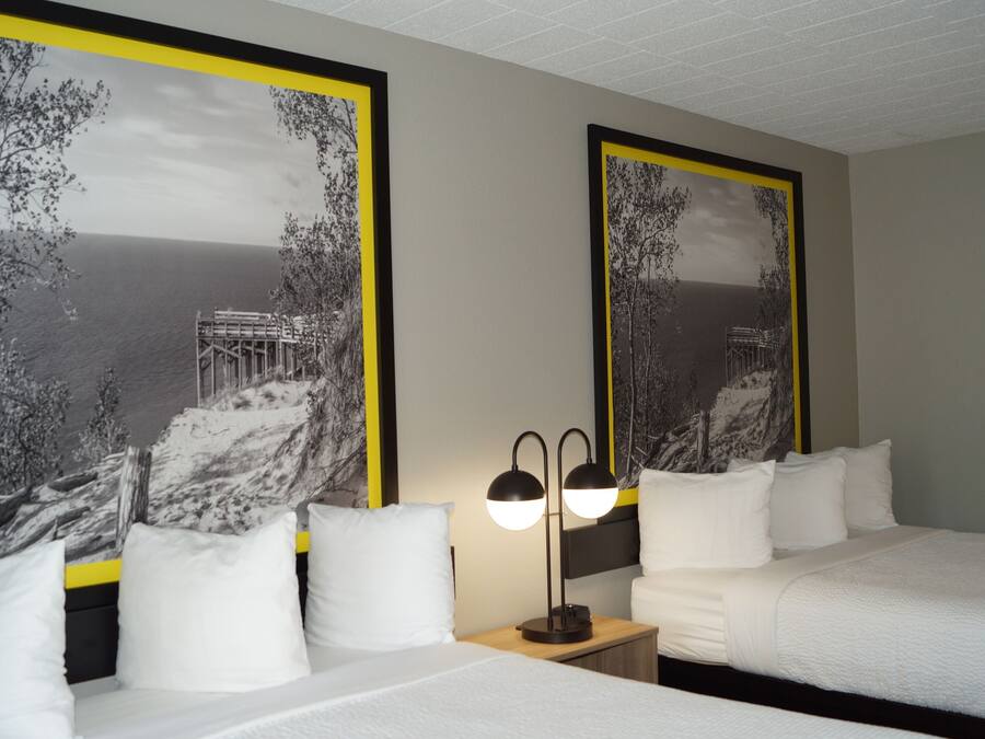 Une chambre d’hôtel avec deux lits, du linge de lit blanc, une table de chevet avec une lampe, et de grandes peintures encadrées en noir et blanc représentant des paysages, accrochées aux murs.