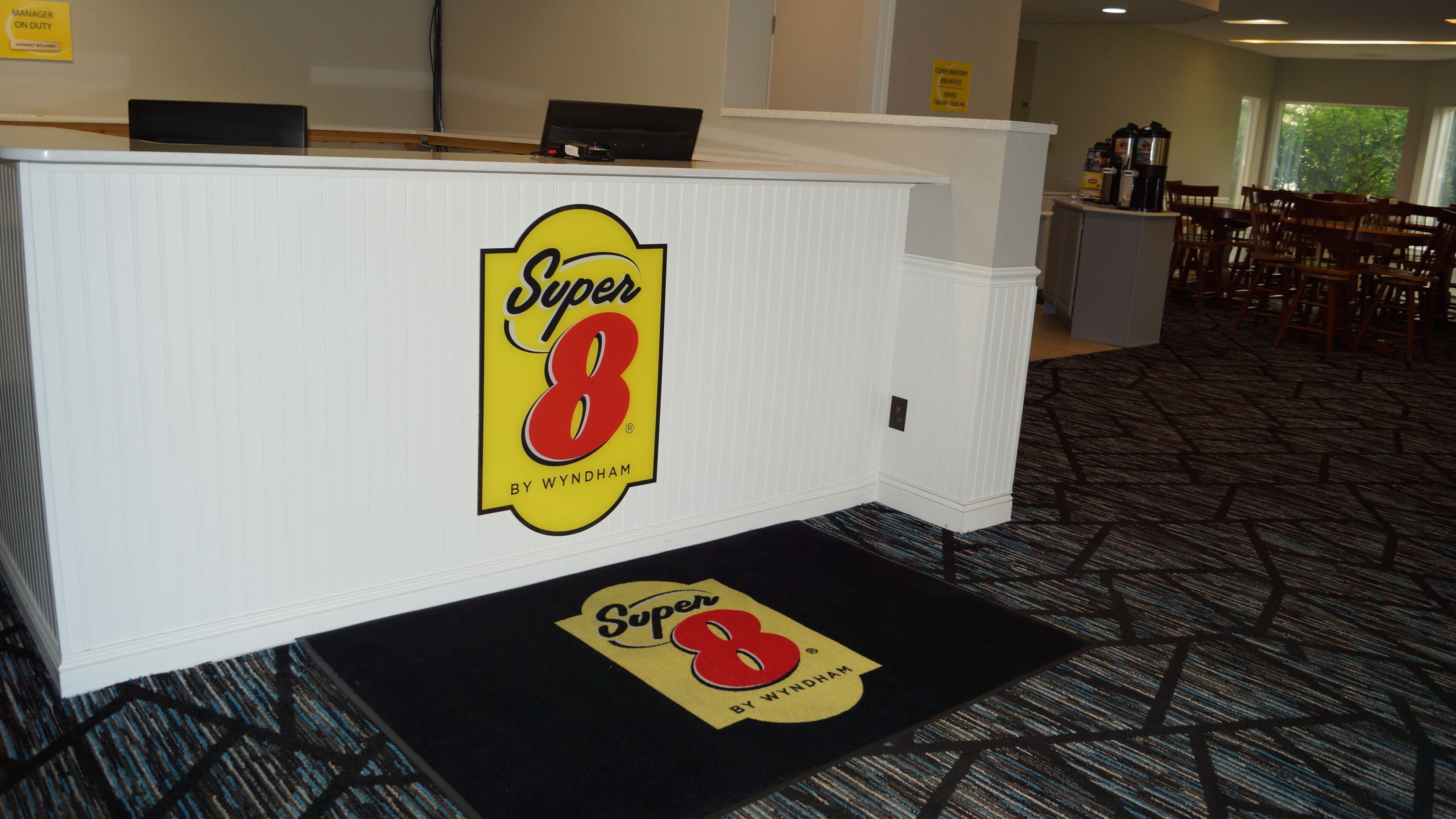 Un comptoir d’accueil orné du logo « Super 8 », un tapis à motifs noir et blanc et un coin repas avec des chaises et des tables en bois en arrière-plan.