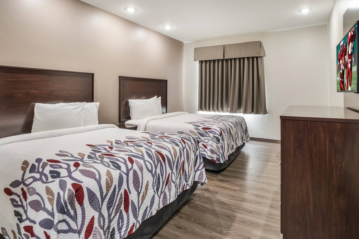 Een hotelkamer met twee bedden, een houten hoofdbord, een groot raam met gordijnen en een flatscreen televisie aan de muur.
