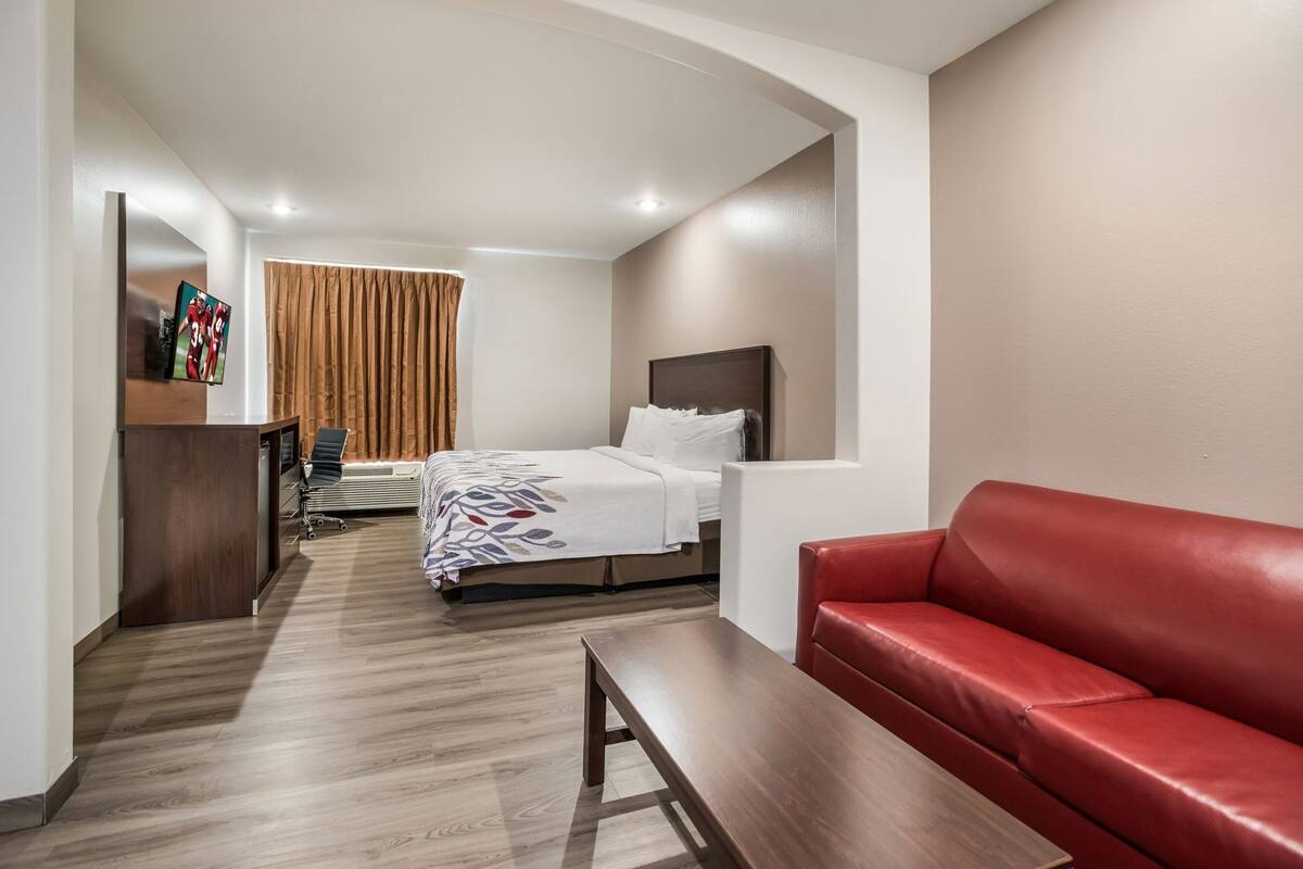 Hotelkamer met een bed, een rode leren bank, een houten bankje, een bureau met stoel en een televisie aan de muur.