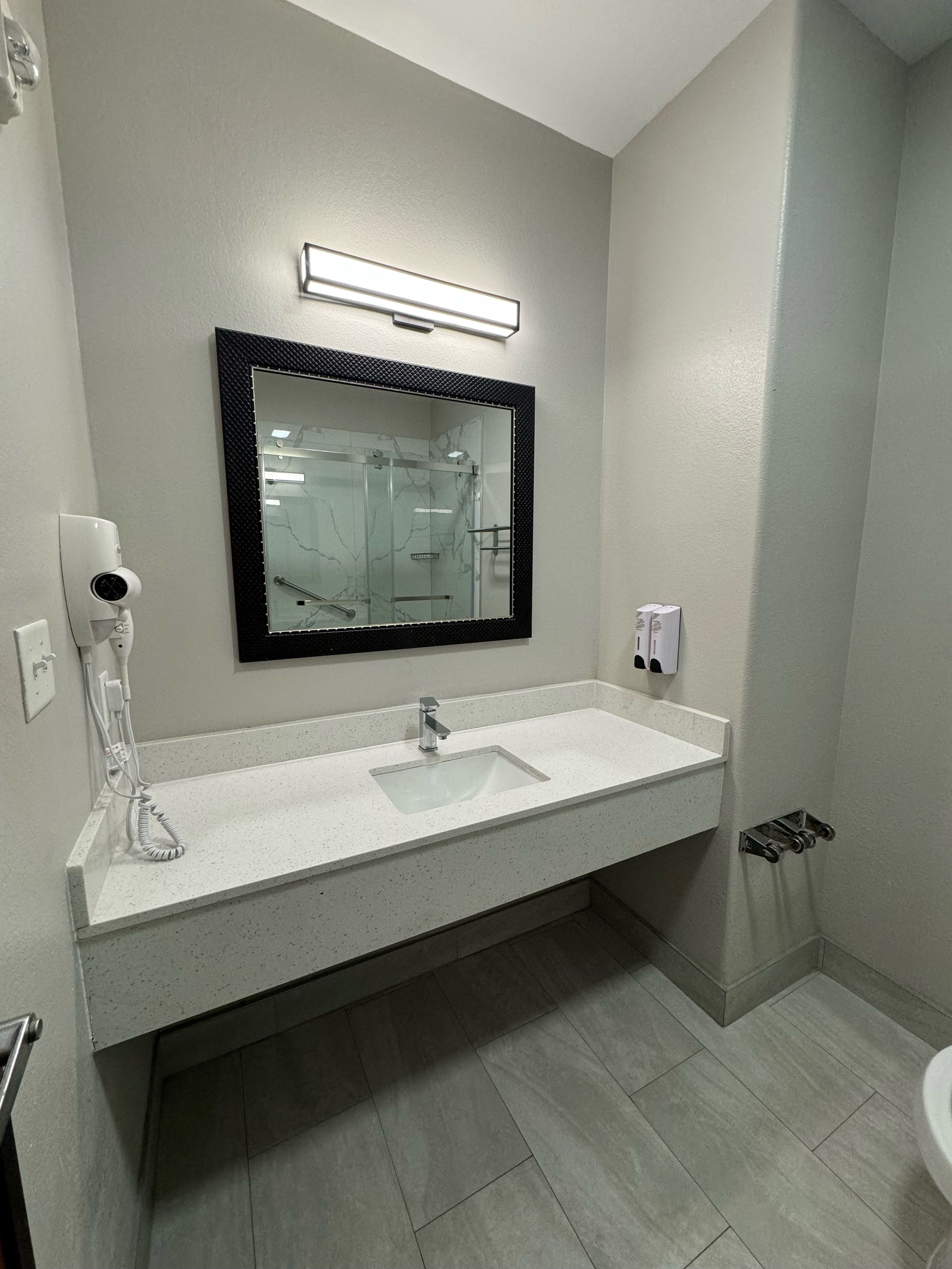 Un baño con un lavamanos blanco, un espejo con marco negro, un secador de pelo de mano y un dispensador de toallas.